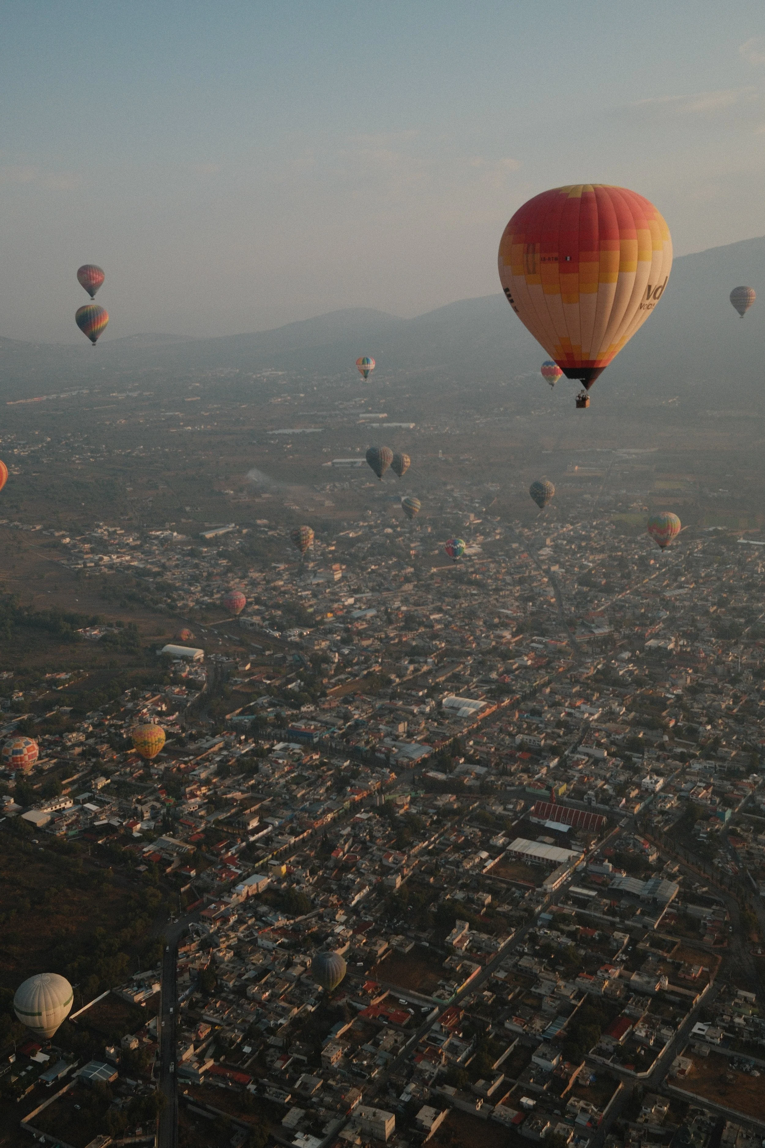 Multiple hot air balloons floating over a city at dawn or dusk, with mountains in the background.