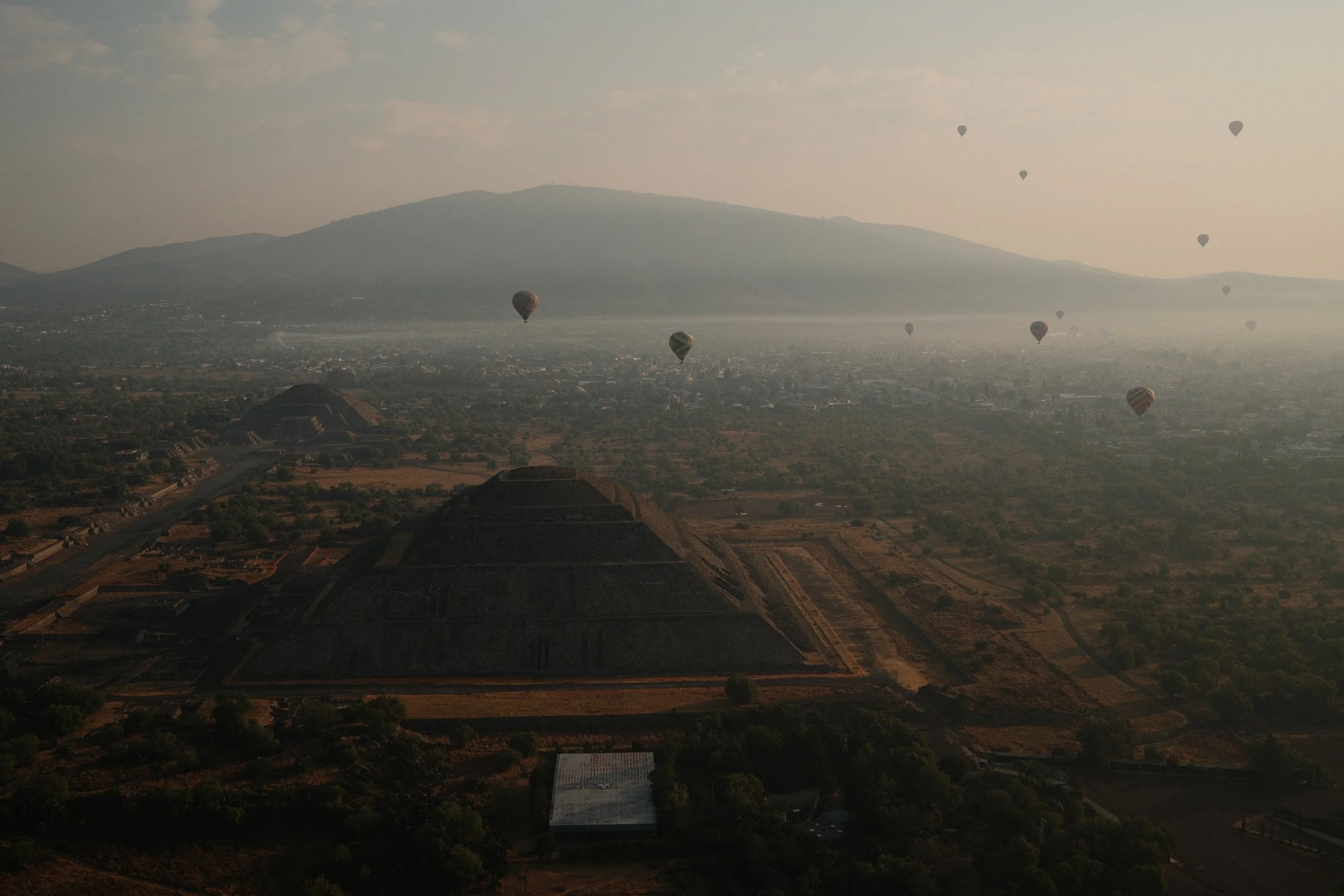 Multiple hot air balloons floating above ancient pyramids in a desert landscape, with misty mountains in the background.