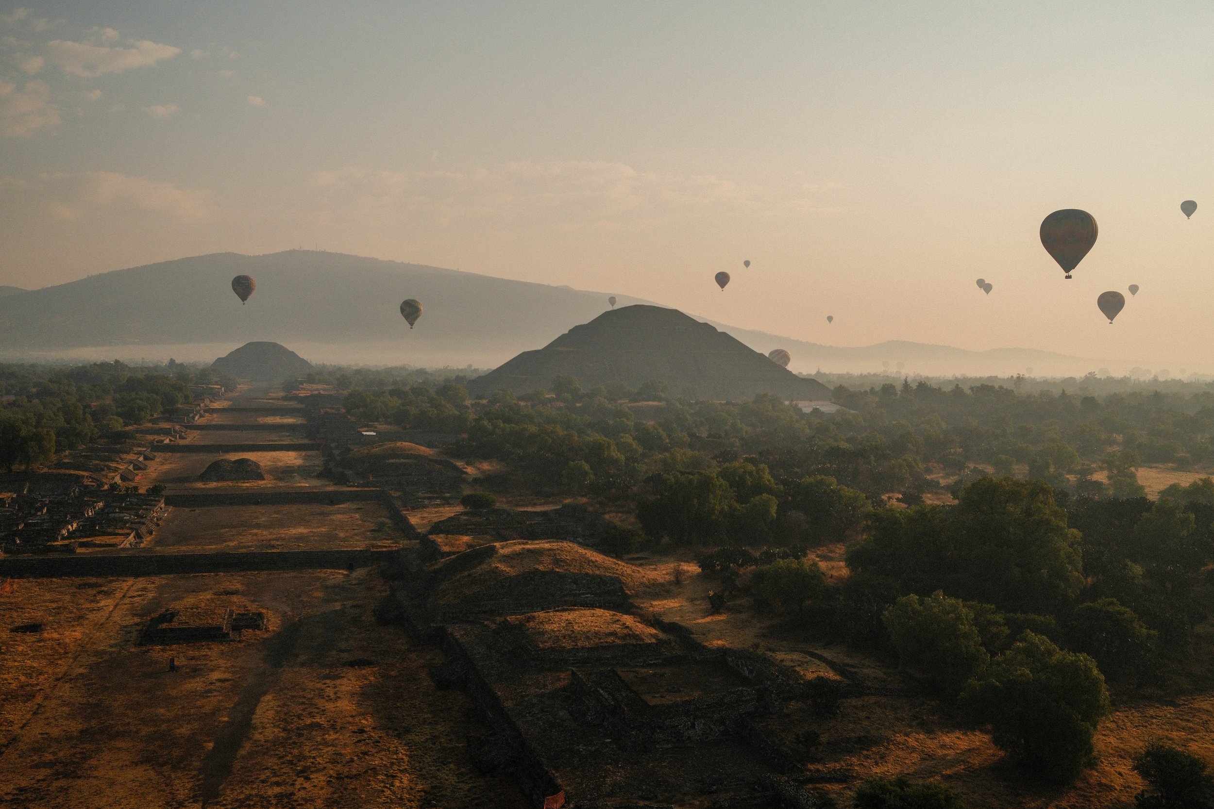 Hot air balloons floating over ancient ruins and lush greenery with mountains in the background during sunrise or sunset.