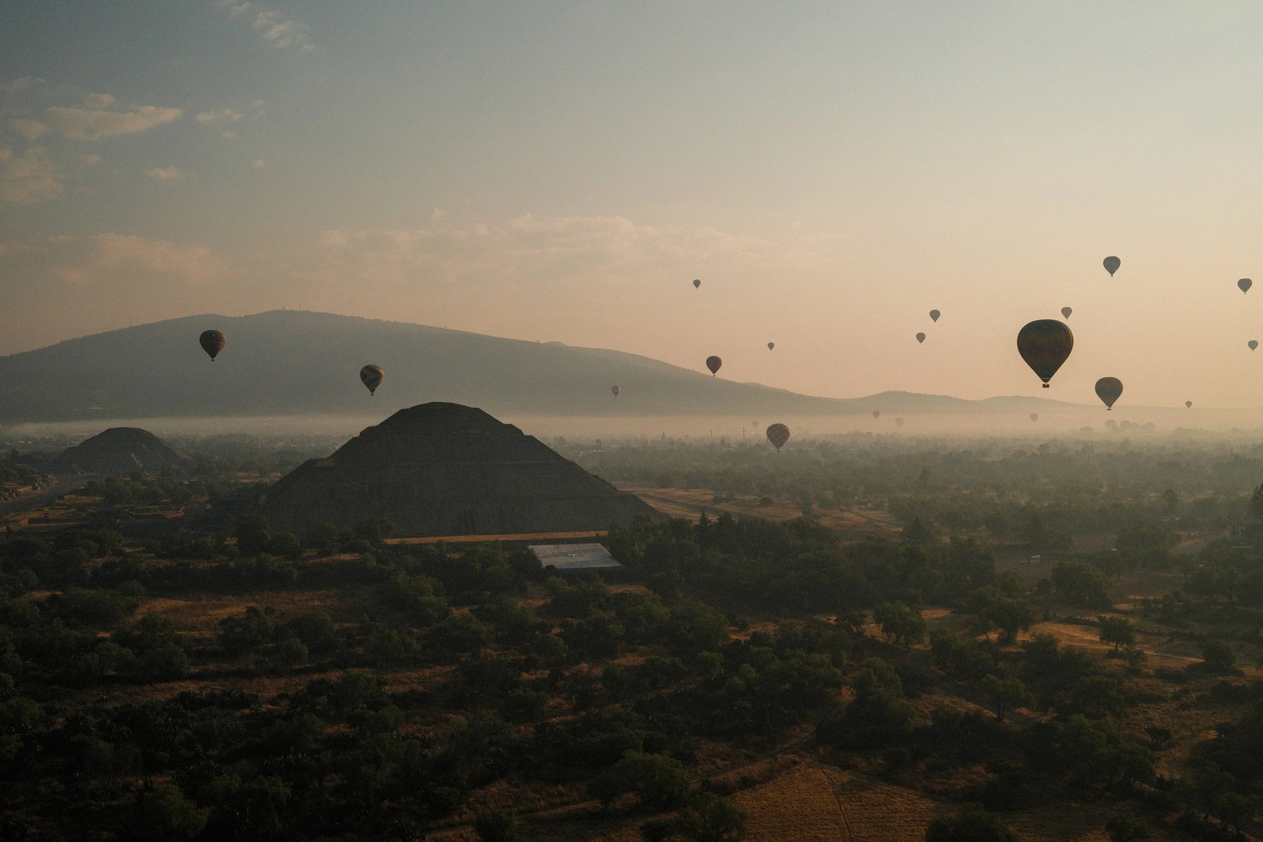 Multiple hot air balloons floating over a landscape with pyramids, trees, and distant mountains during sunrise or sunset.
