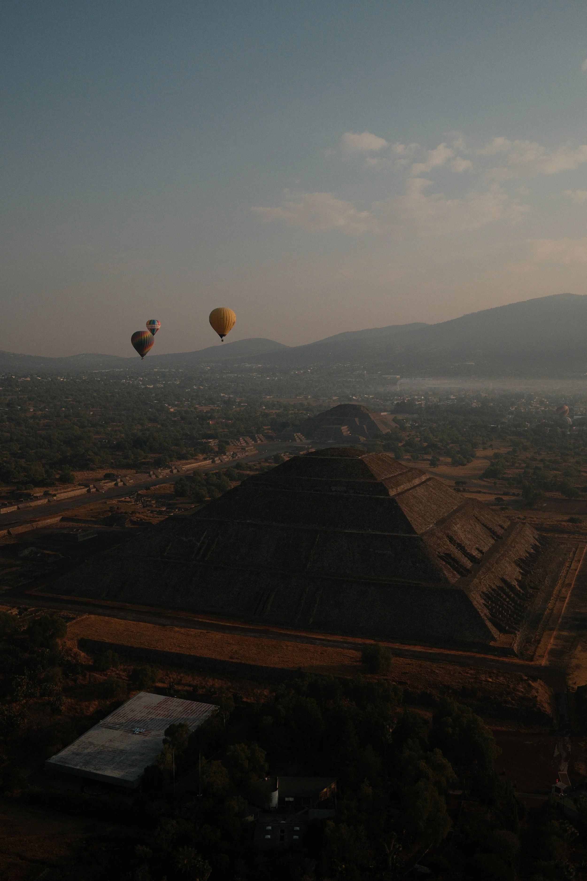 Hot air balloons floating above the ancient pyramids of Mexico at sunrise or sunset with mountains in the background.