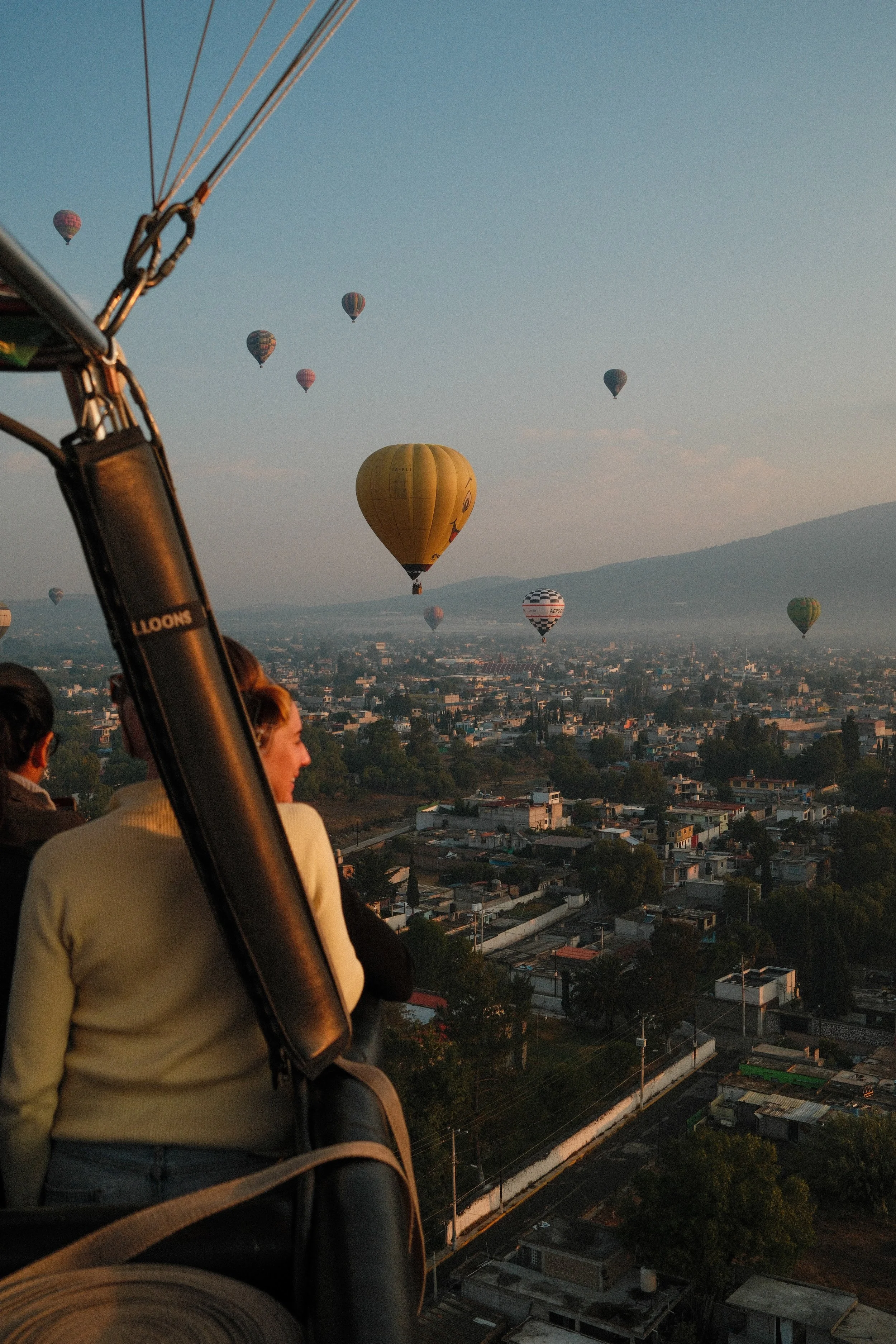 View from a hot air balloon showing several other hot air balloons floating in the sky over a city with mountains in the background during sunset.
