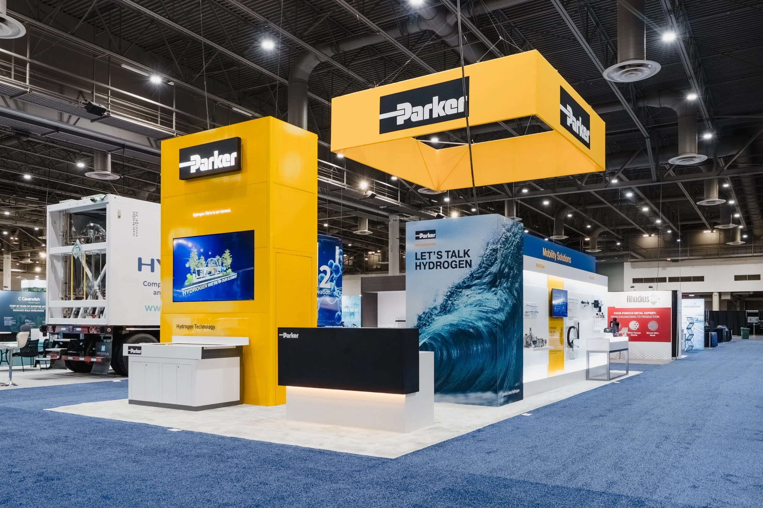 Trade show booth for Parker with yellow and blue branding, featuring displays about hydrogen technology and mobility solutions, inside a large exhibition hall with other booths and equipment in the background.