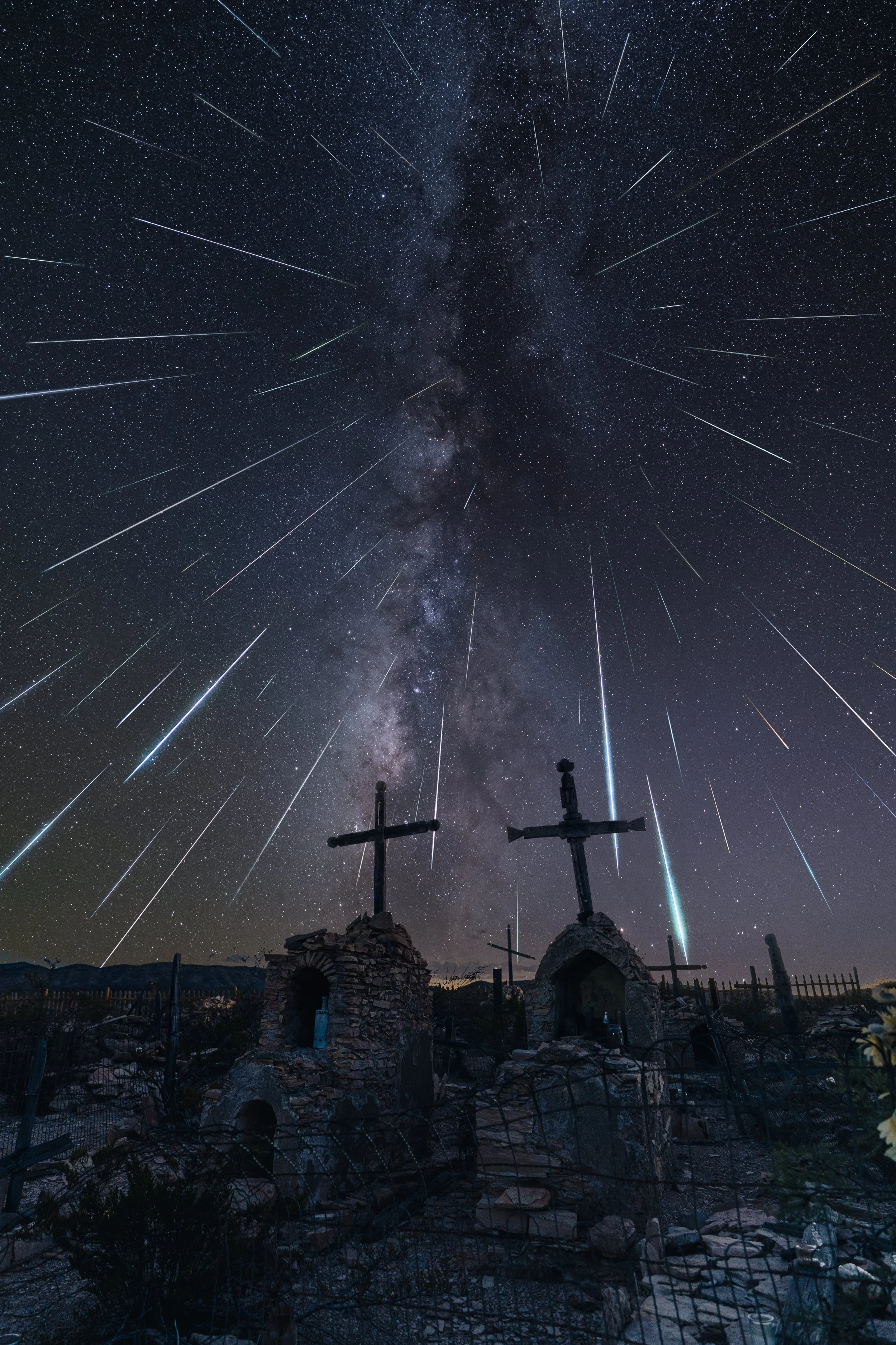 Night sky filled with stars and streaking shooting stars above a landscape with stone structures and crosses.