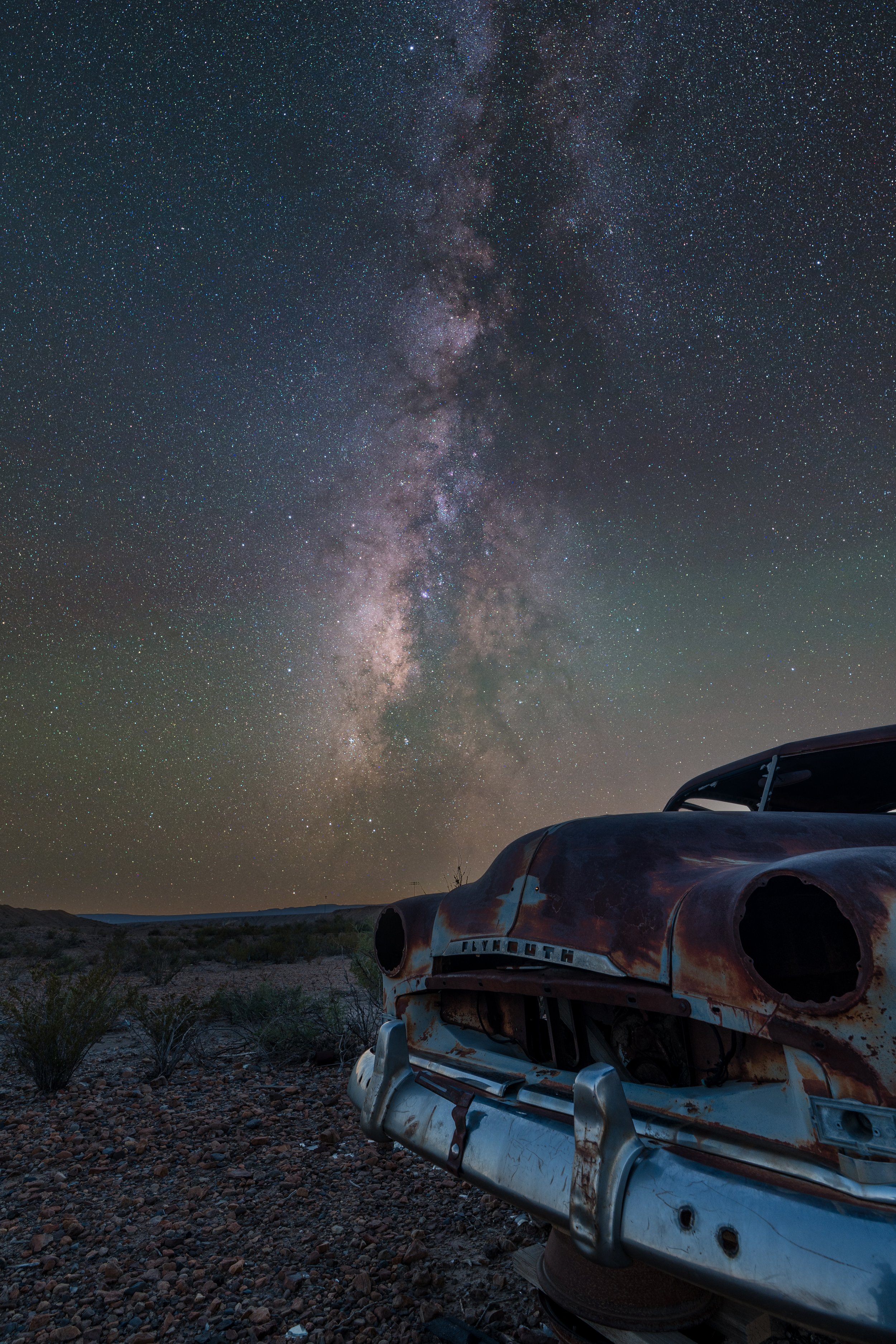A rusty, abandoned Plymouth car in a desert landscape under a clear night sky filled with stars and the Milky Way galaxy.