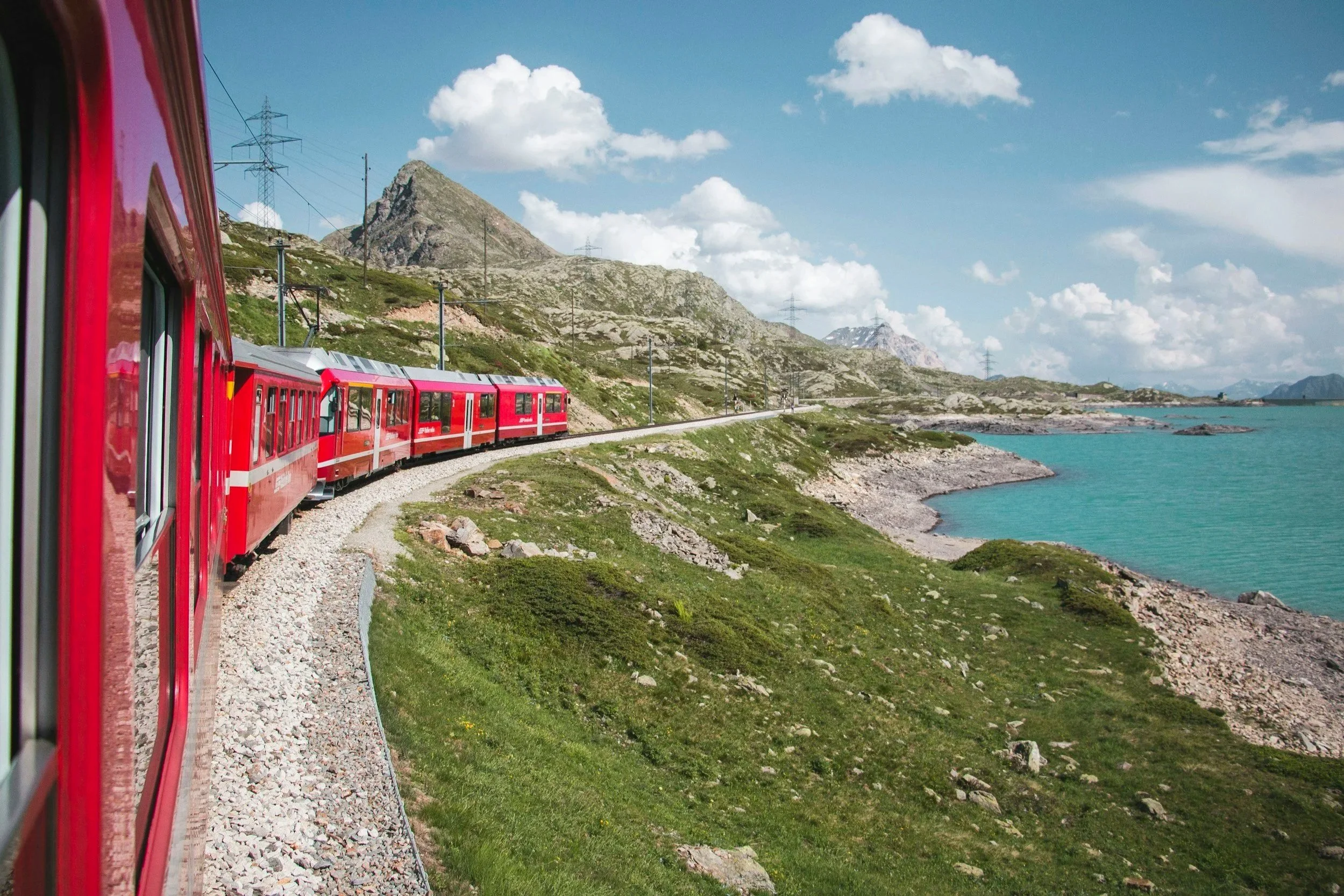 Red Bernina Express train curving along a mountain track next to a bright turquoise Alpine lake under a cloudy sky.