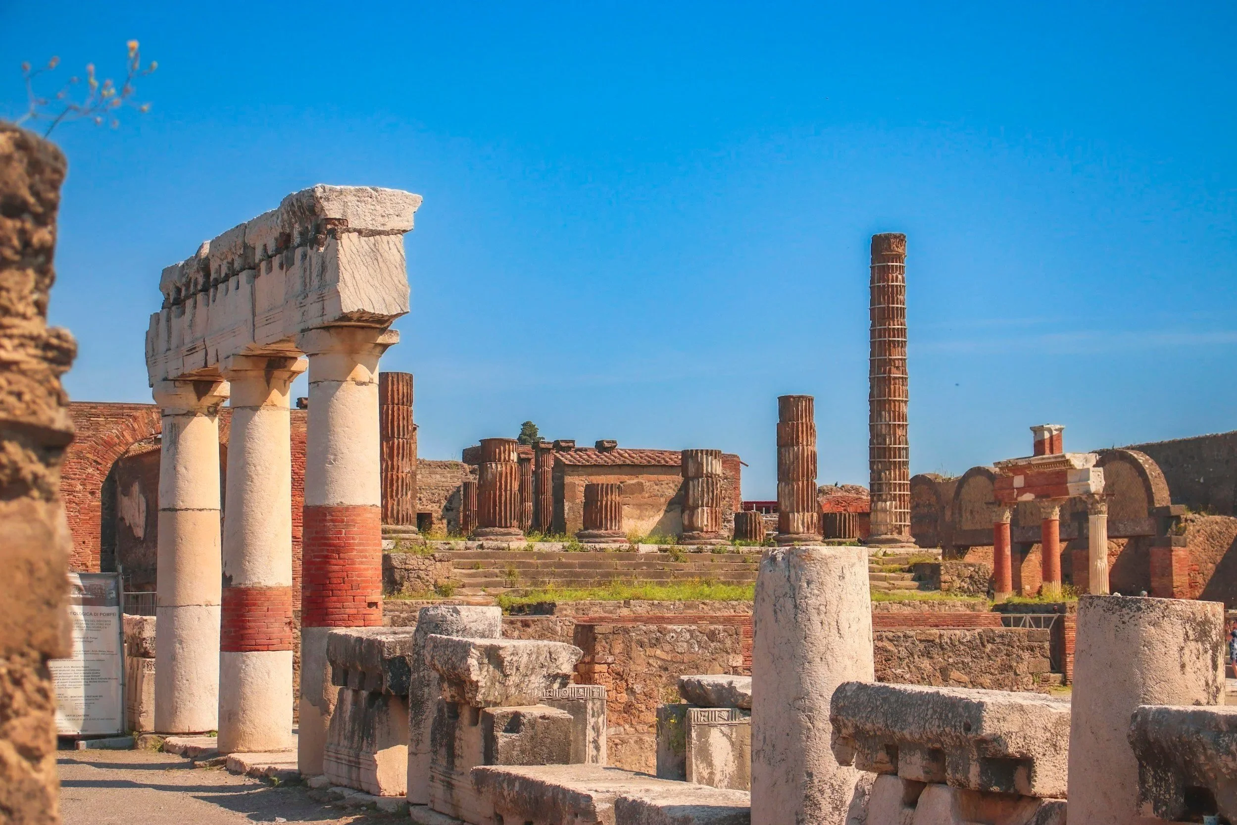 Ancient Roman stone columns and brick ruins at the Basilica in the Pompeii archaeological site under a clear blue sky.