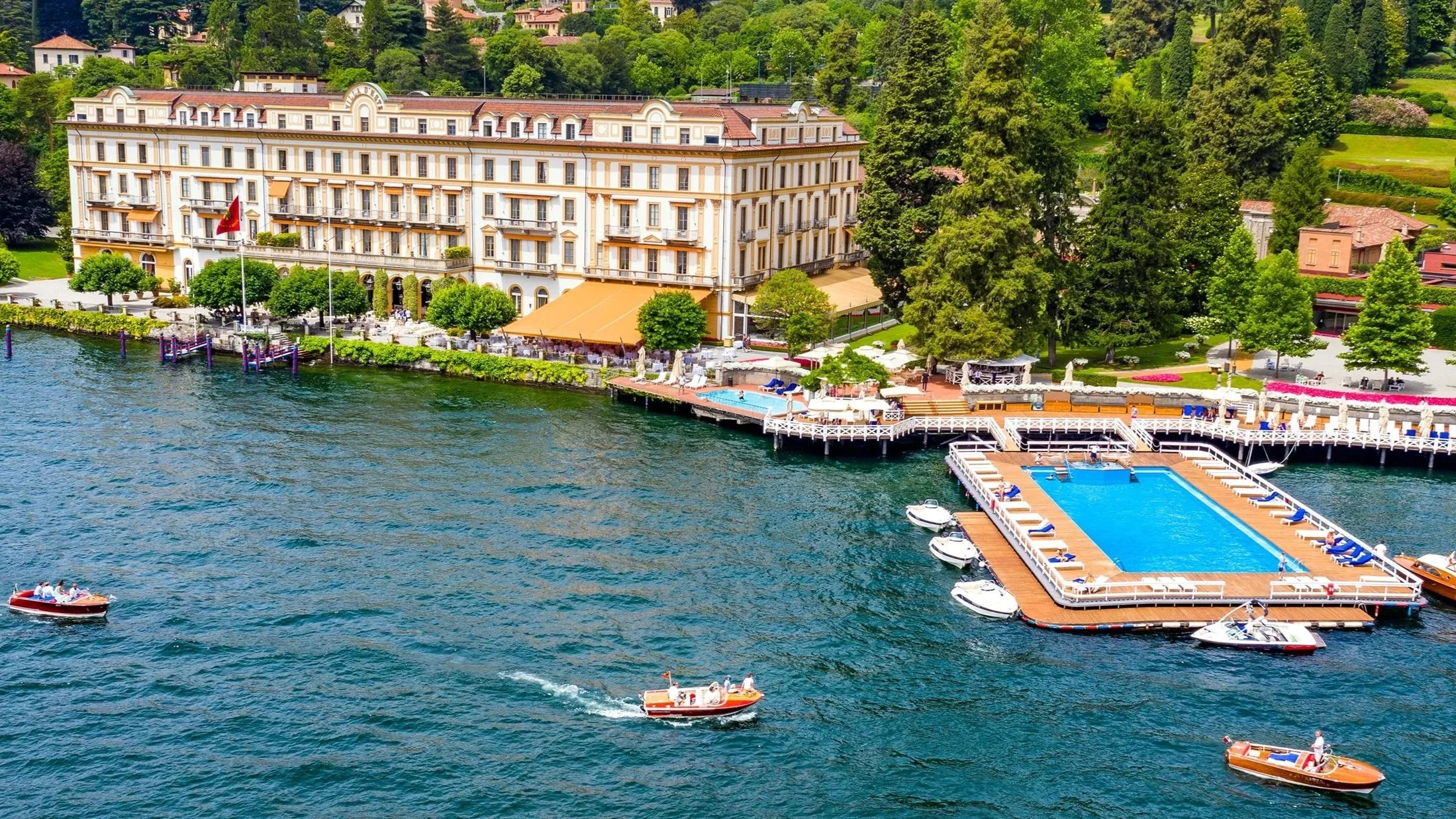 Aerial waterfront view of the historic Villa d’Este luxury hotel and resort with its famous floating pool on Lake Como surrounded by boats.