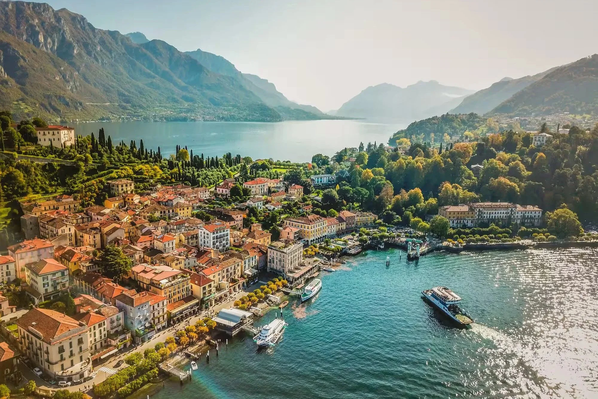High-angle panoramic view of Bellagio village with colorful lakeside buildings, ferries on Lake Como, and surrounding mountains.