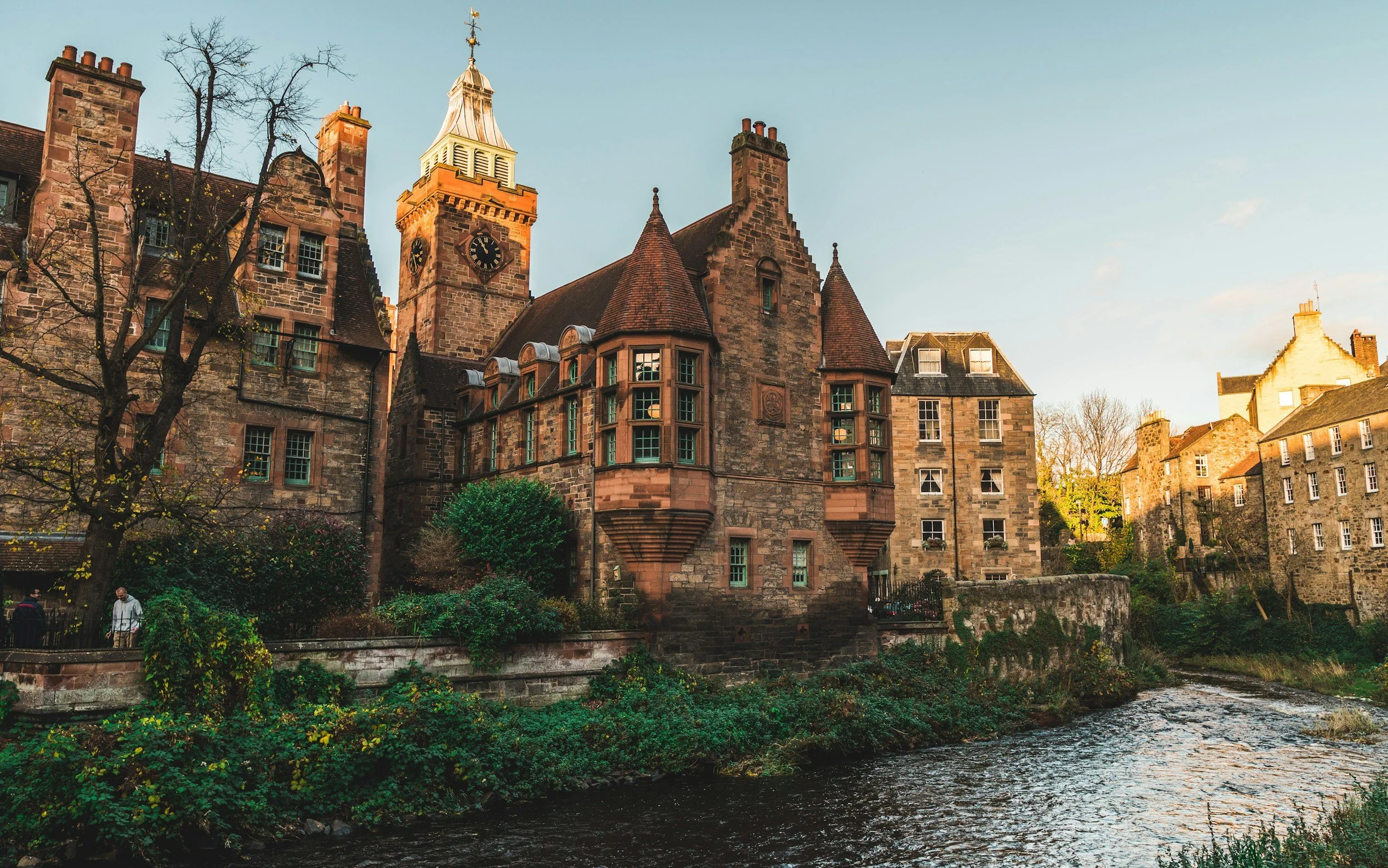 Historic stone buildings in Edinburgh, Scotland with spires and a clock tower line a serene river. The warm sunlight casts a golden glow, creating a tranquil, timeless scene.
