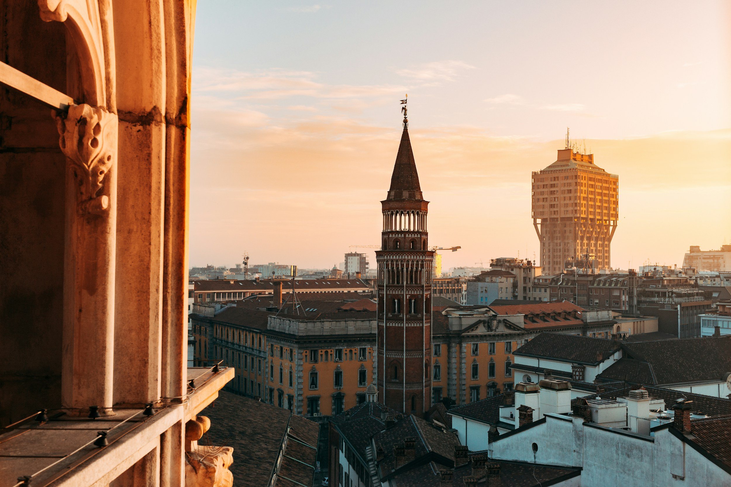 Elevated rooftop view of Milan at sunset showing the brick bell tower of San Gottardo in Corte framed against the unique profile of Torre Velasca.