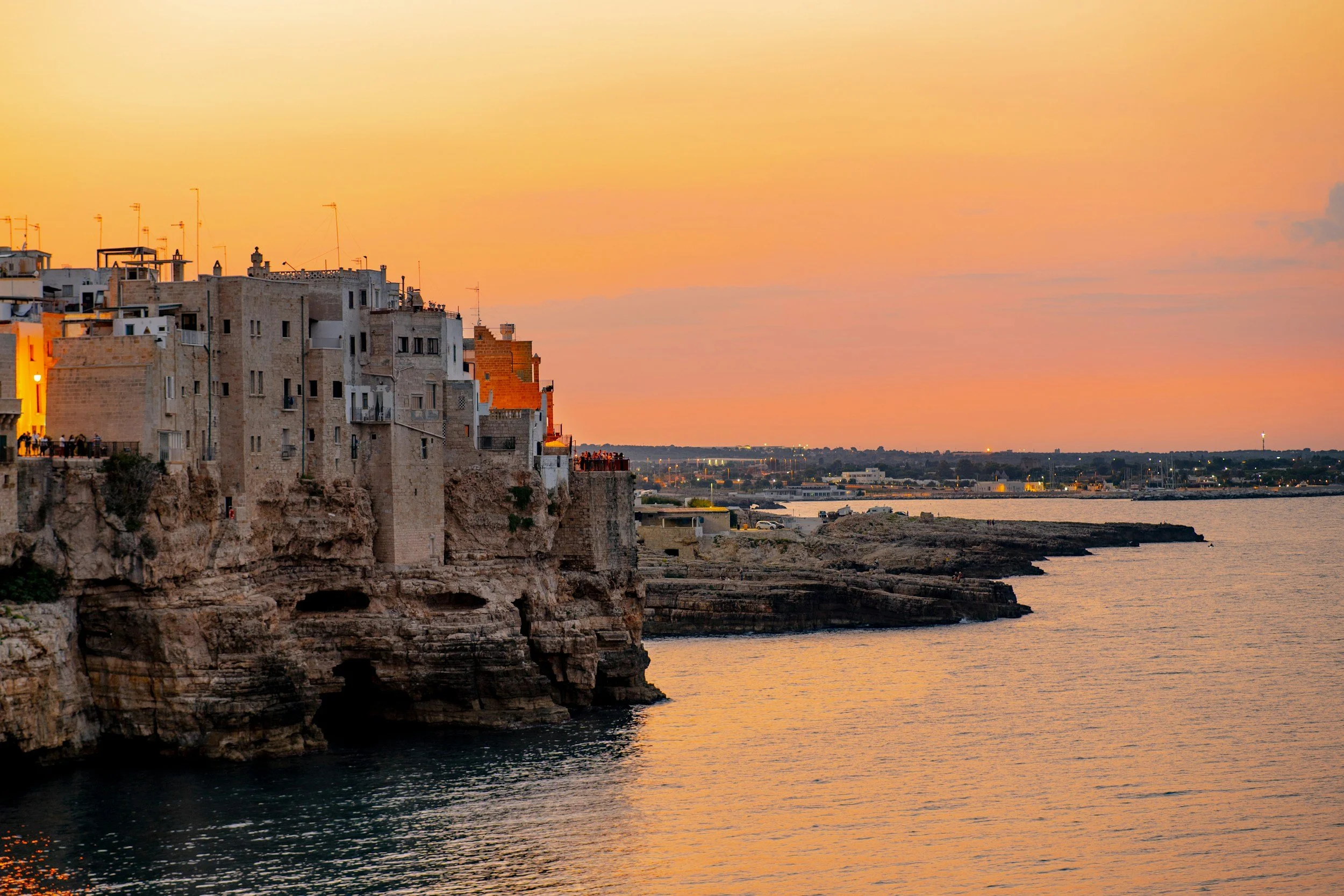 Warm sunset glow behind the cliffside whitewashed buildings and sea caves of historic Polignano a Mare overlooking the calm Adriatic waters in Puglia, Italy.