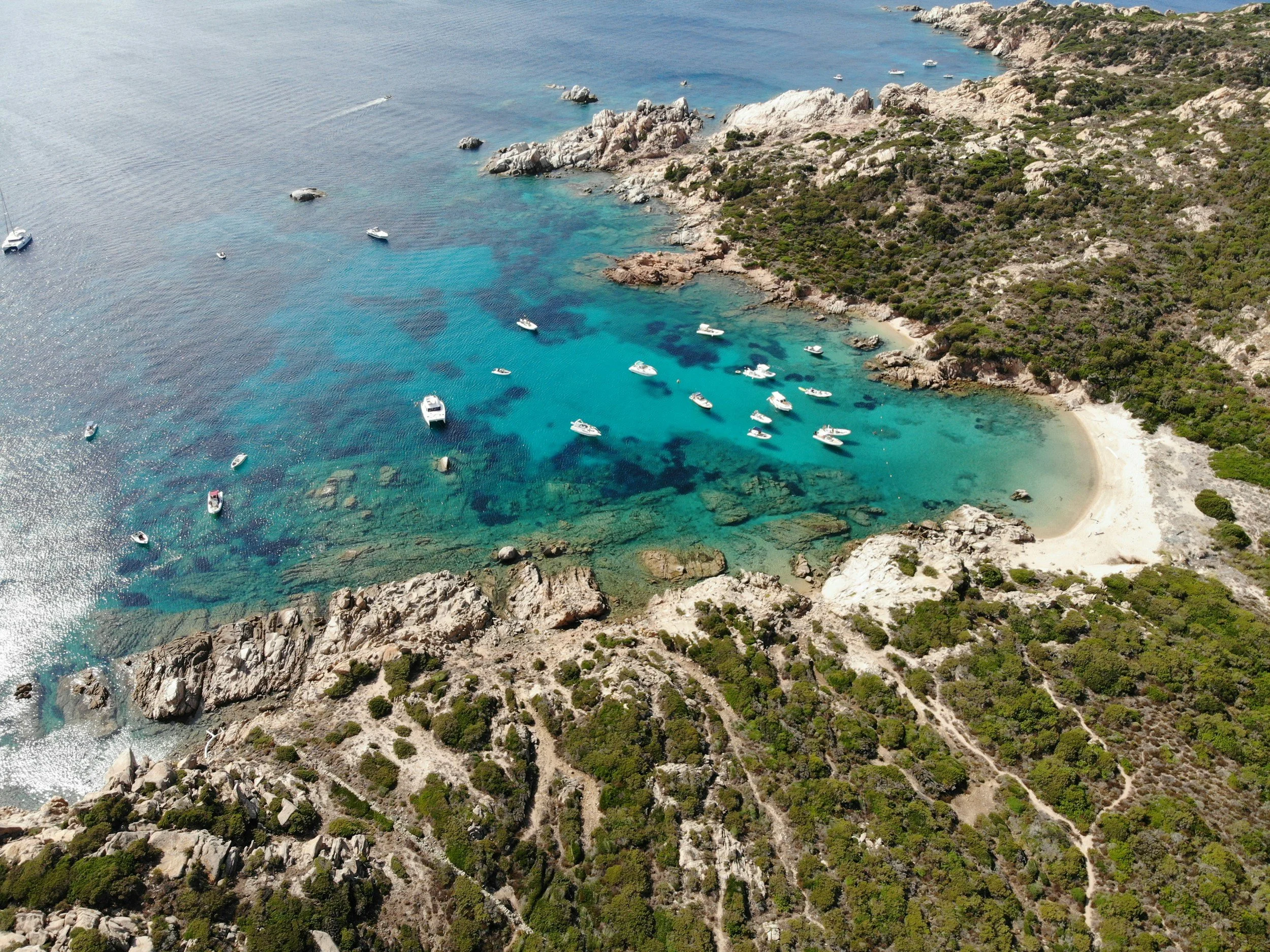 Aerial drone view of small boats anchored in a secluded turquoise cove surrounded by rugged granite rock and green Mediterranean maquis in Sardinia.