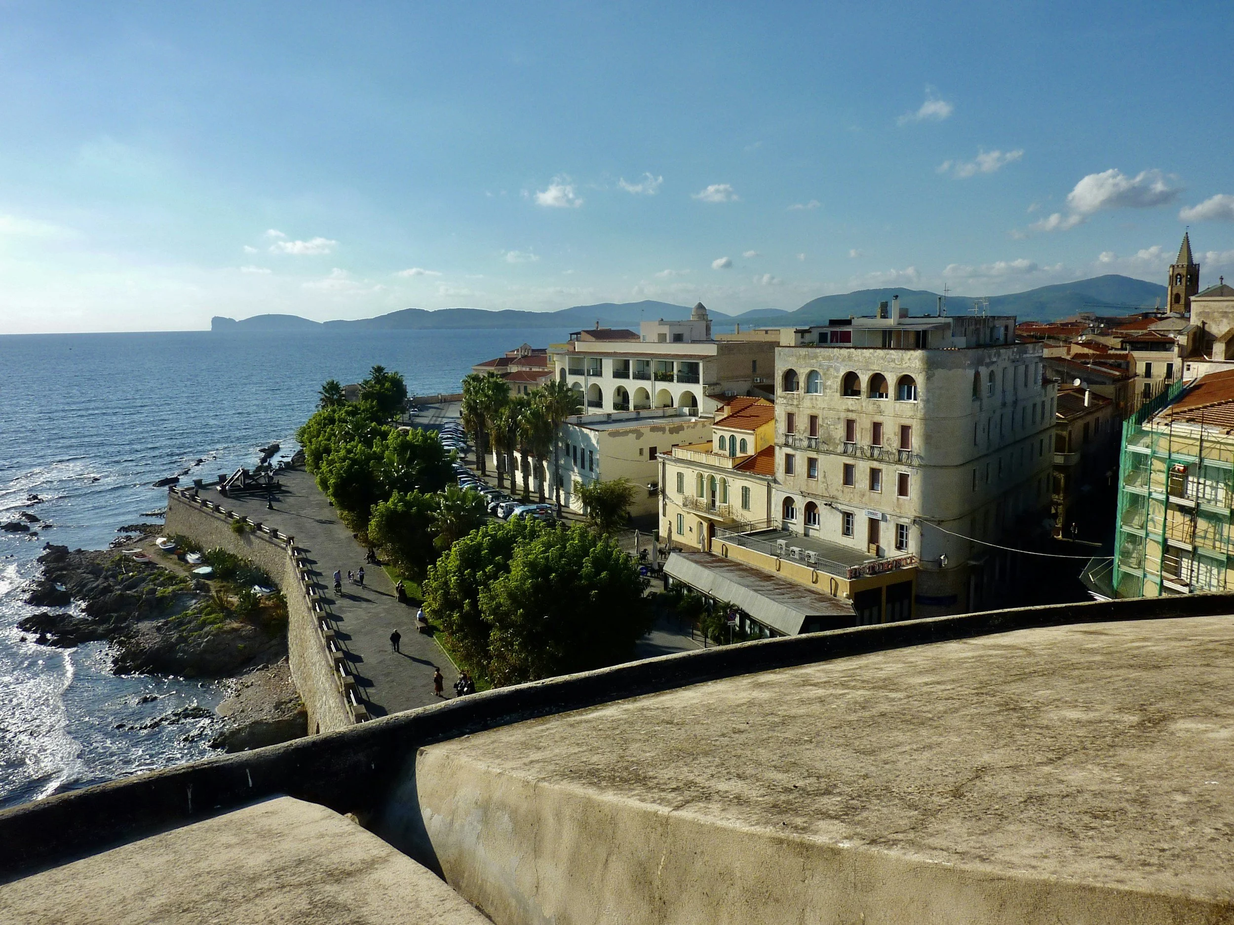 Elevated panoramic view of Alghero's historic sea bastions, promenade lined with trees, and the Mediterranean coastline stretching towards Capo Caccia.