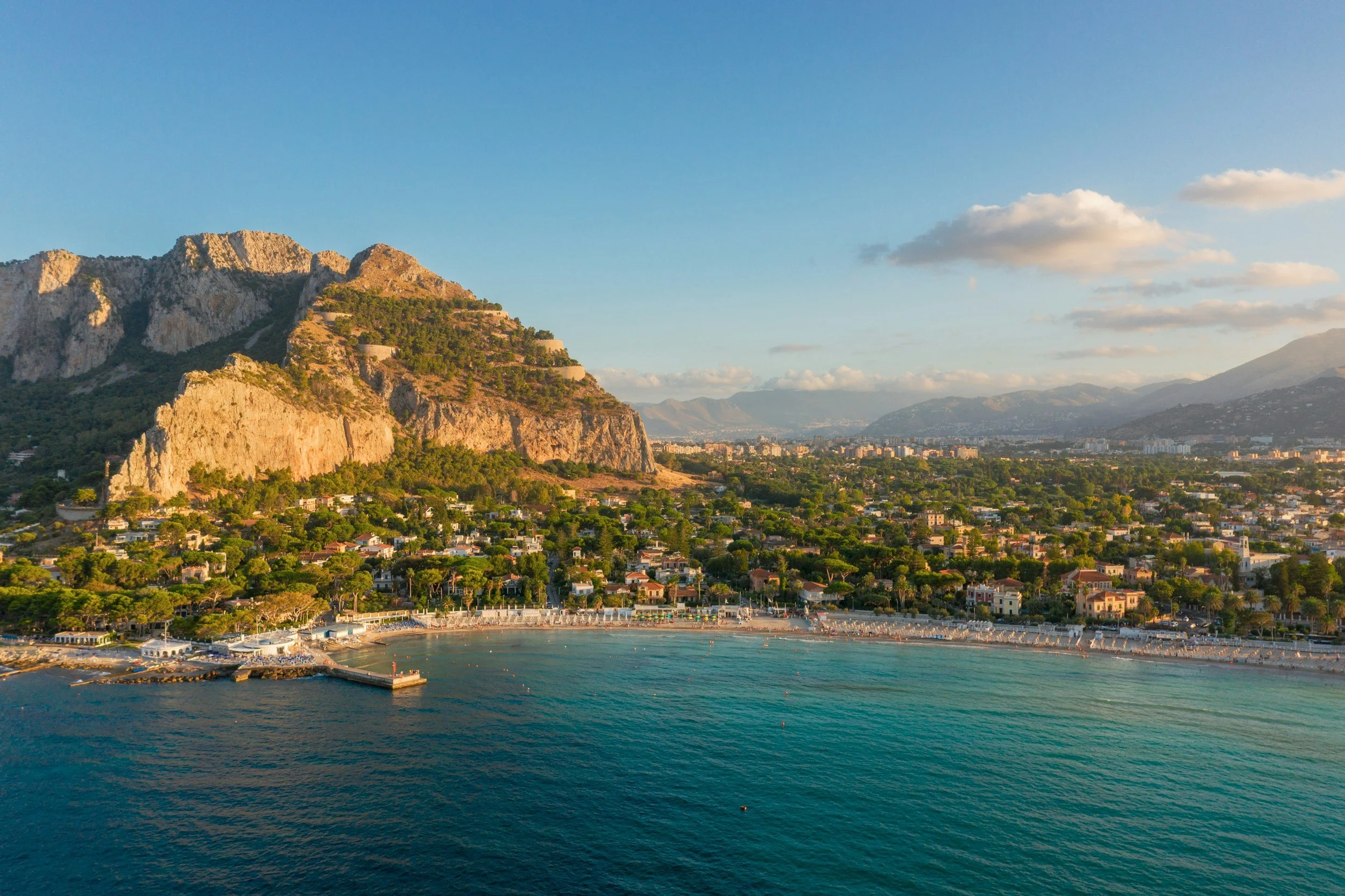 High-angle aerial shot of Mondello Beach bay and clear blue sea framed by the dramatic limestone cliffs of Monte Pellegrino in Palermo, Sicily.