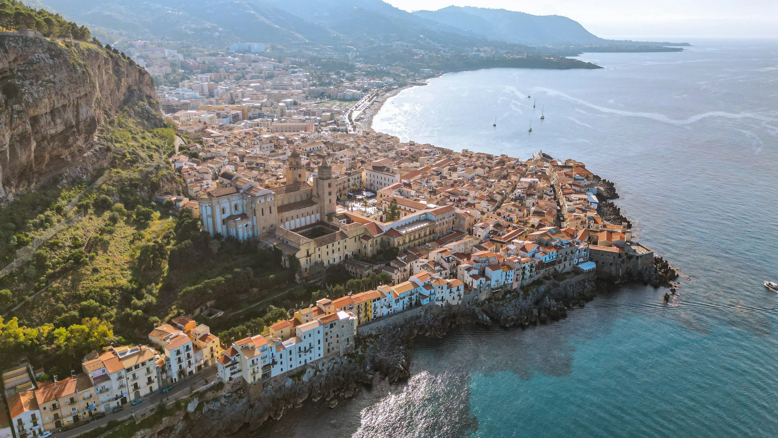 High aerial view of Cefalù’s historic old town nestled between the cliff of La Rocca and the blue Mediterranean Sea, showing the grand Norman Cathedral and waterfront buildings.