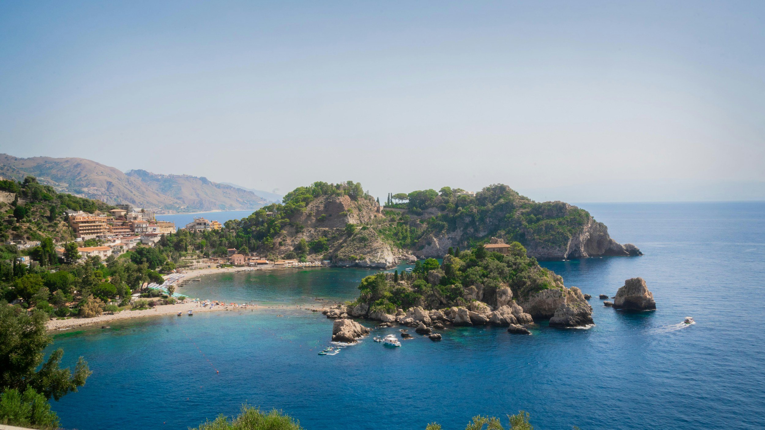 High-angle panoramic view of Isola Bella nature reserve connected to the pebble beach by a narrow tombolo sandbar in the turquoise Ionian Sea below Taormina.