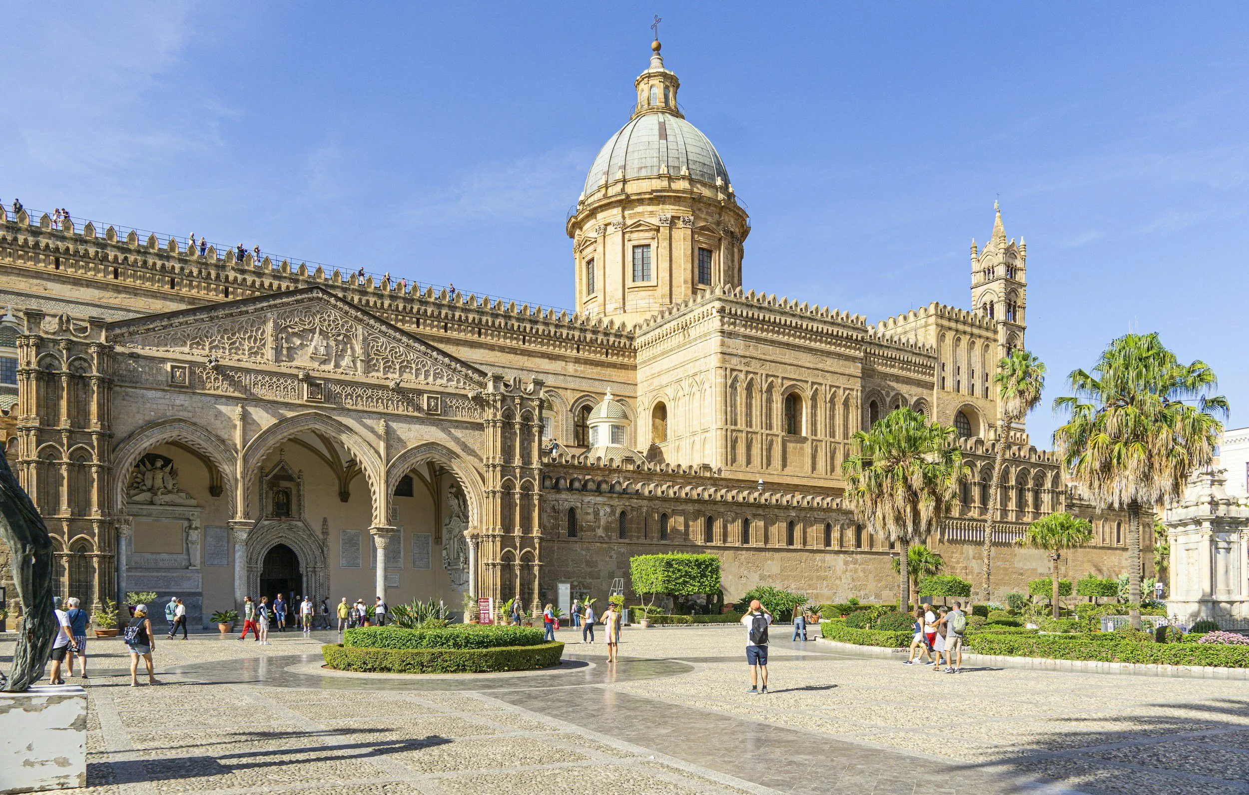 Sunlit exterior façade and grand dome of the Palermo Cathedral featuring ornate Arab-Norman arches and palm trees in the surrounding piazza.