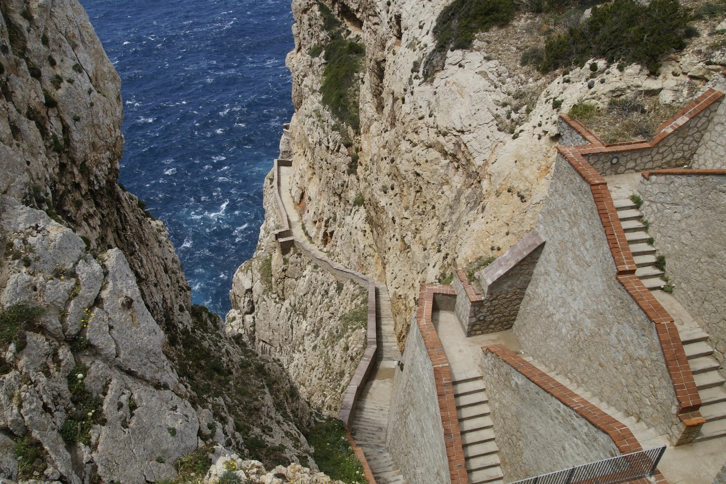 High-angle view of the Escala del Cabirol, a dramatic stone staircase zigzagging down steep limestone cliffs above the deep blue sea at Capo Caccia, Sardinia.
