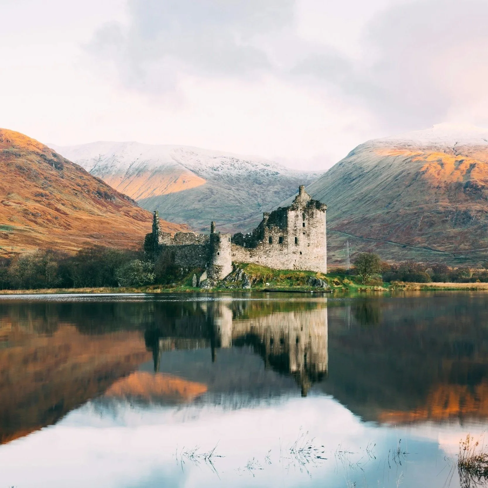 A serene lakeside castle ruin in Scotland reflects in calm water, surrounded by snow-capped mountains under a cloudy sky. The scene evokes tranquility and timelessness.