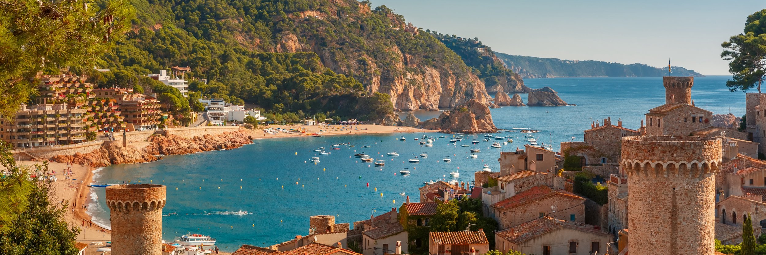 View of Tossa de Mar on the Costa Brava, Spain, showing the medieval Vila Vella fortress walls and towers overlooking the crescent-shaped bay, beach, and steep, wooded cliffs.