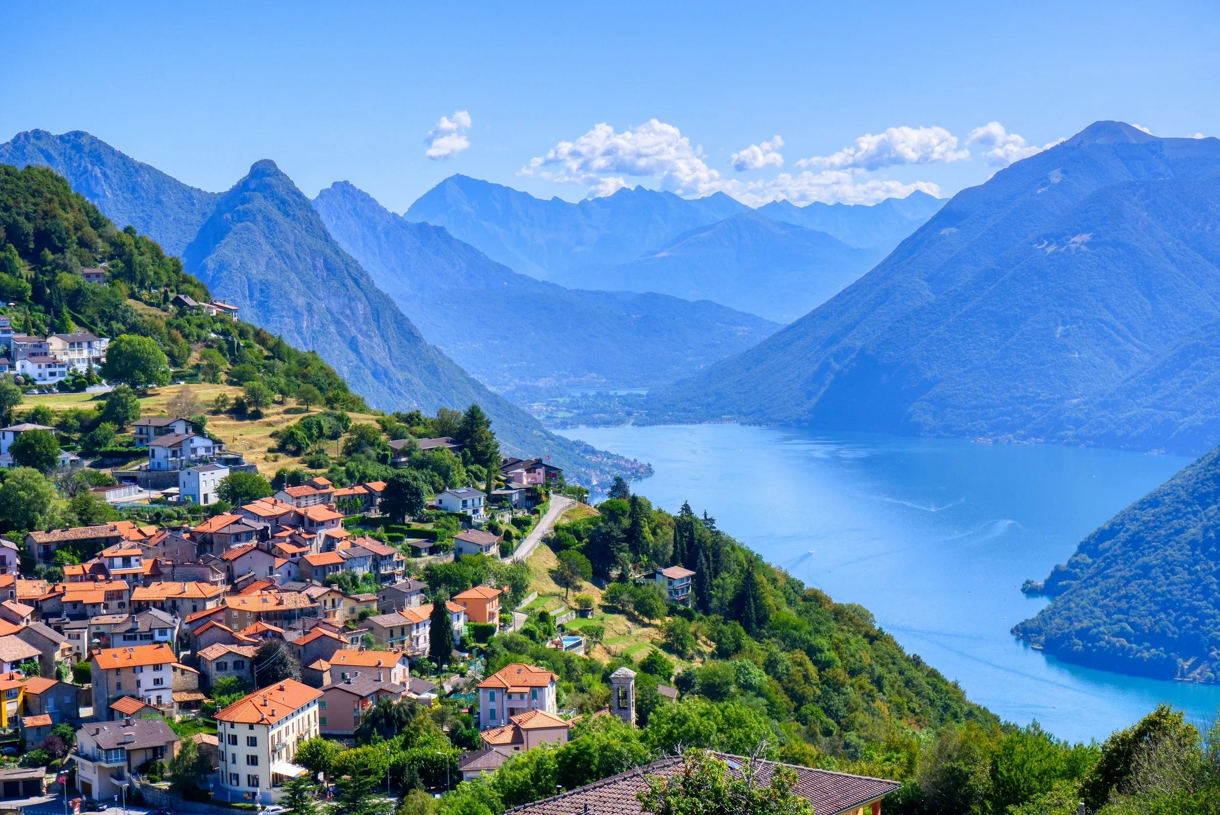 A picturesque village of Lugano with red-roofed houses nestled on a lush hillside overlooks a serene blue lake, framed by majestic, hazy mountains under a clear sky.