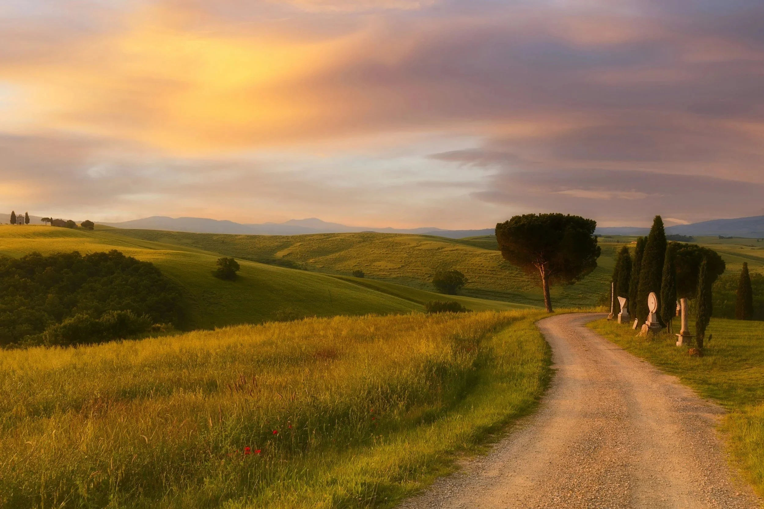A winding gravel country road curving through rolling green hills and cypress trees under a glowing sunset sky in the Italian countryside