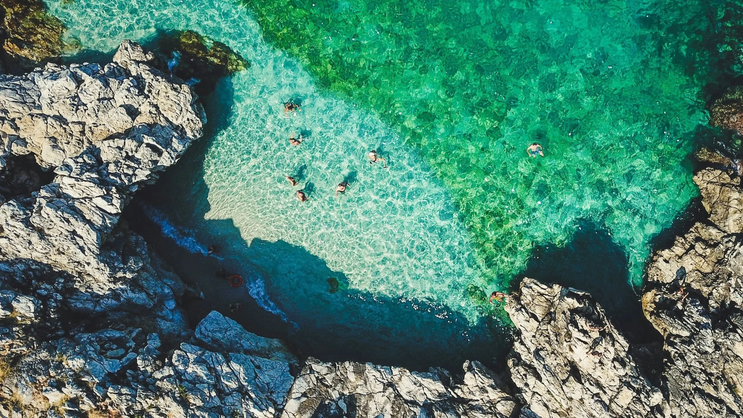 Aerial top-down view of swimmers in crystal-clear turquoise waters framed by limestone cliffs at Zingaro Nature Reserve in Sicily.