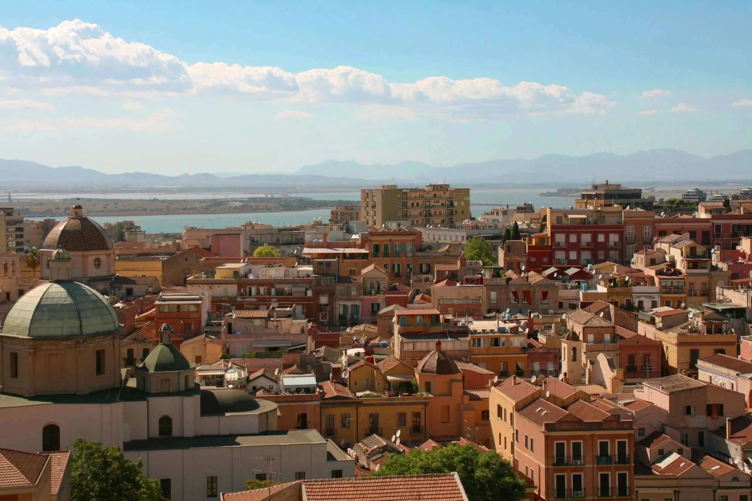 High-angle panoramic view of Cagliari’s historic Old Town rooftops, terracotta domes, and terracotta-tiled buildings overlooking the distant lagoon and mountains in southern Sardinia.