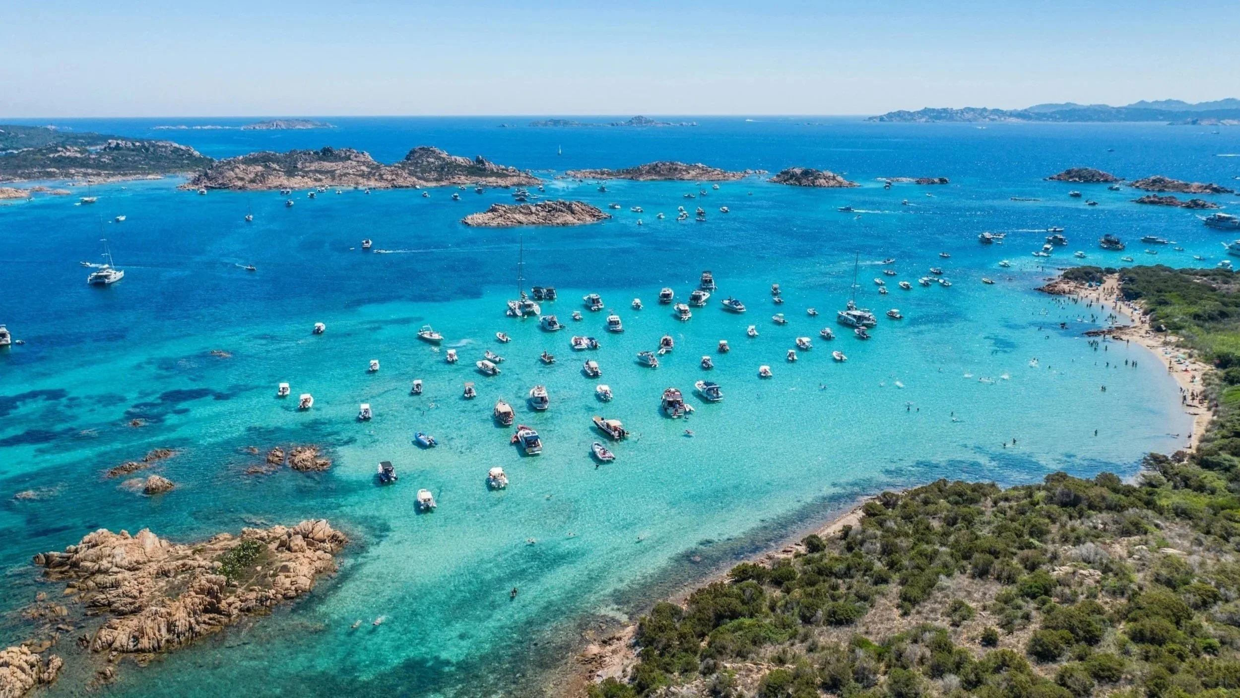 High-angle aerial view of luxury boats and yachts anchored in clear turquoise lagoon waters surrounded by rocky islets in La Maddalena Archipelago, Sardinia.