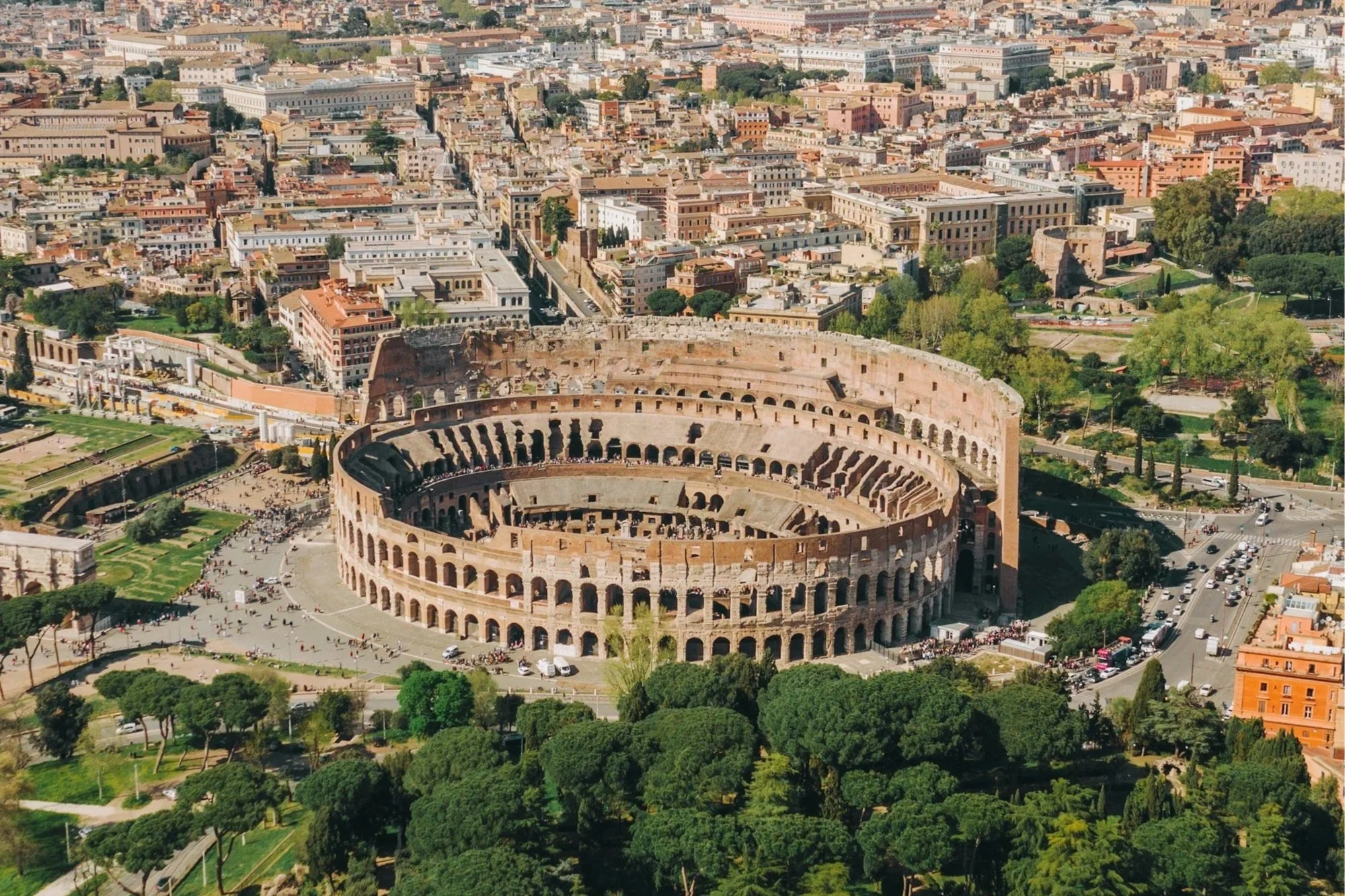 Aerial view of the ancient Colosseum surrounded by green trees, modern streets, and the historic skyline of Rome, Italy.
