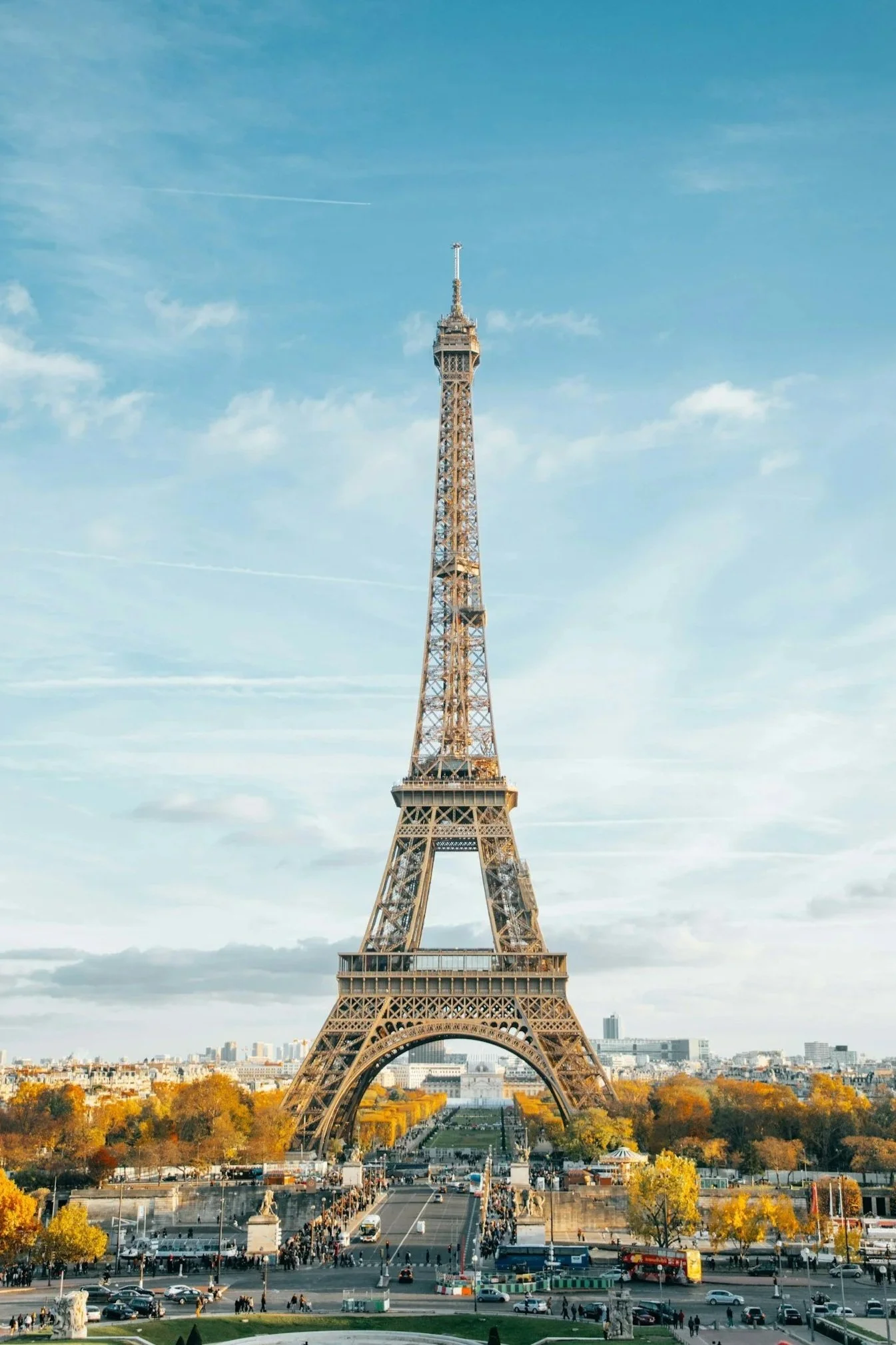 The Eiffel Tower in Paris, France under a blue sky with some clouds and contrails, surrounded by trees, buildings, and a busy street with cars and people.