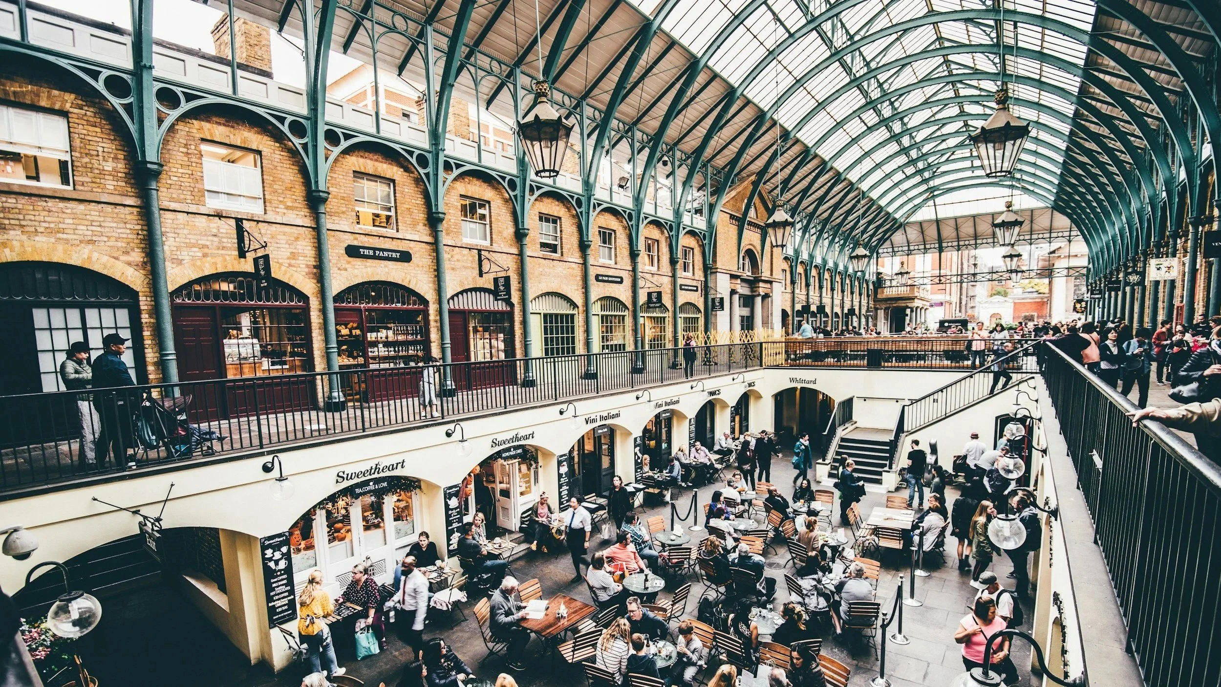 The bustling Covent Garden, an indoor market scene with a tall arched glass roof, iron framework, and brick walls. People are dining and shopping, creating a lively atmosphere.