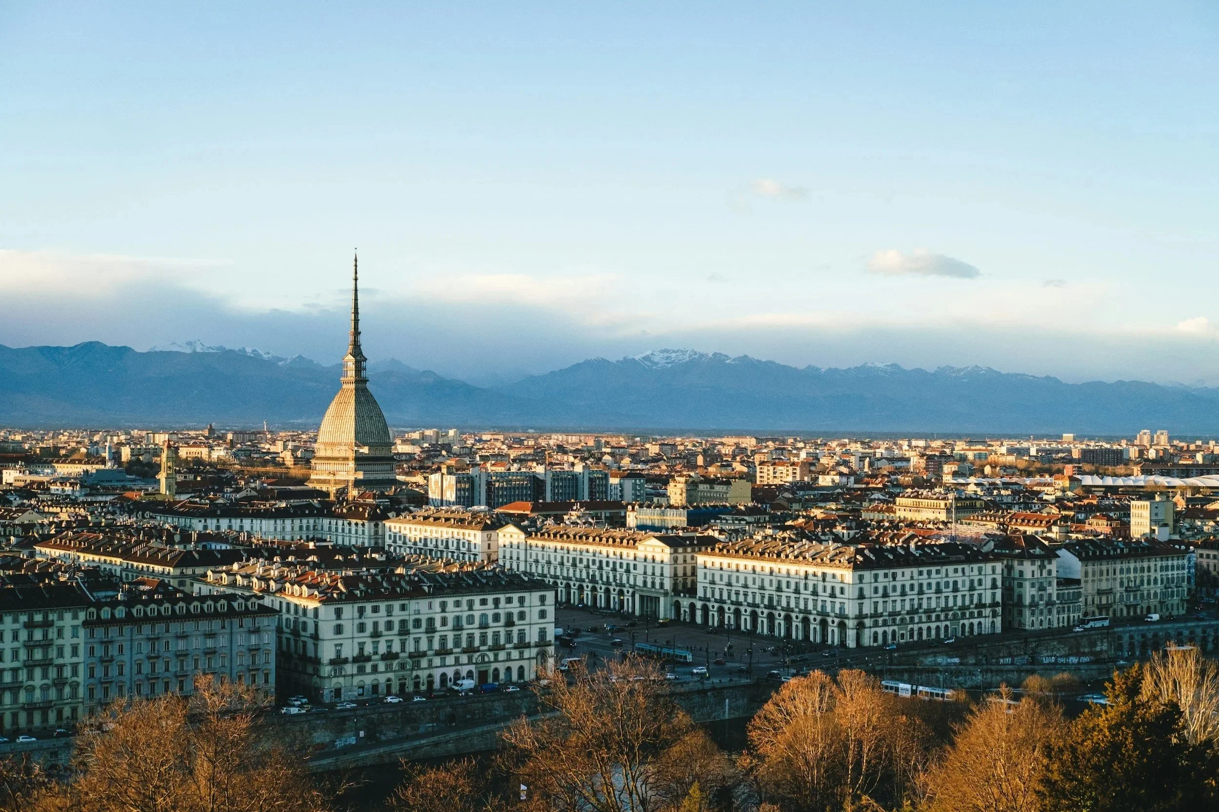 Panoramic skyline view of Turin, Italy featuring the iconic Mole Antonelliana spire with snow-capped Alps in the background.
