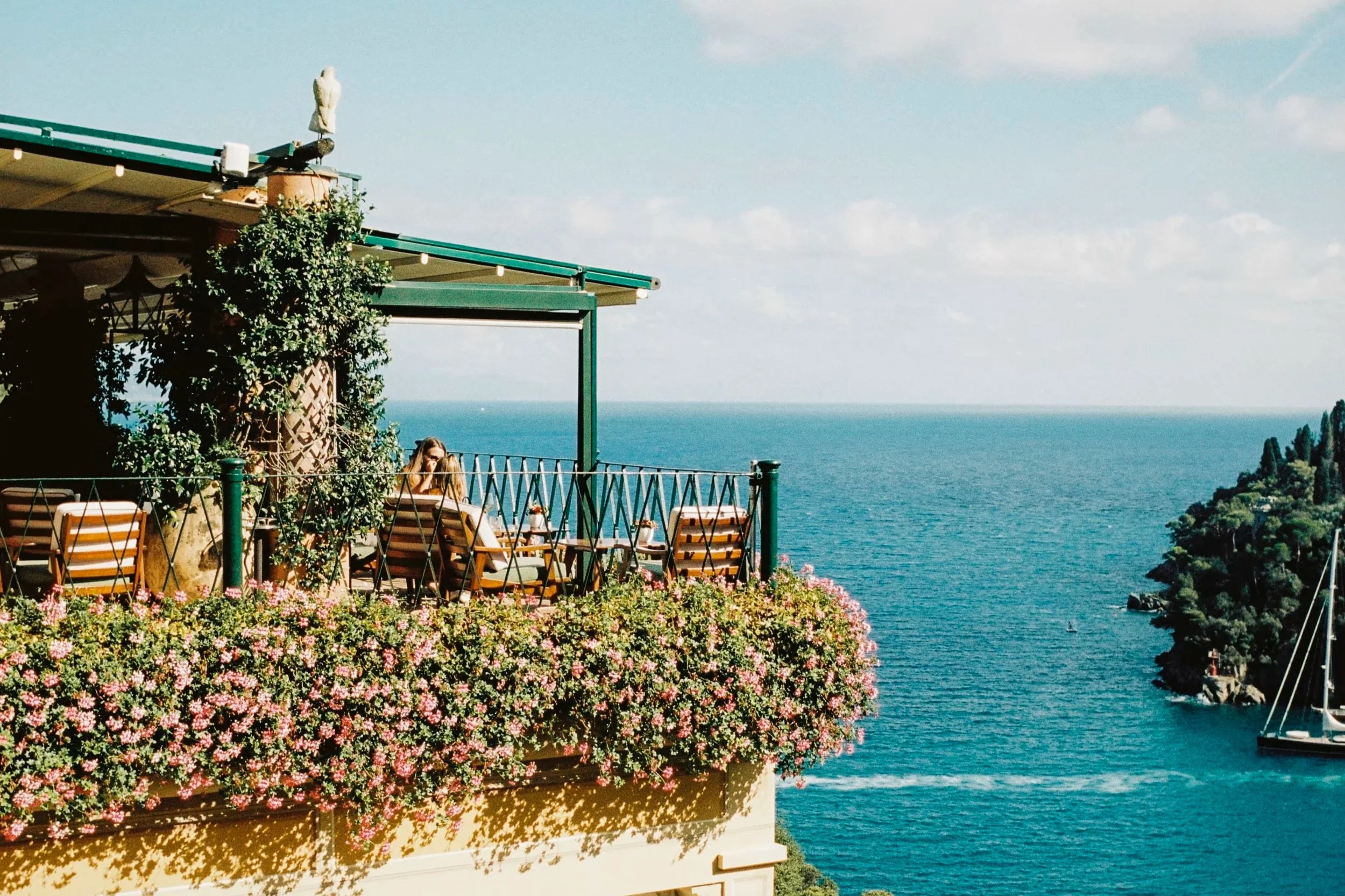 A flower-draped terrace overlooking the deep blue sea and lush peninsula in Italy.
