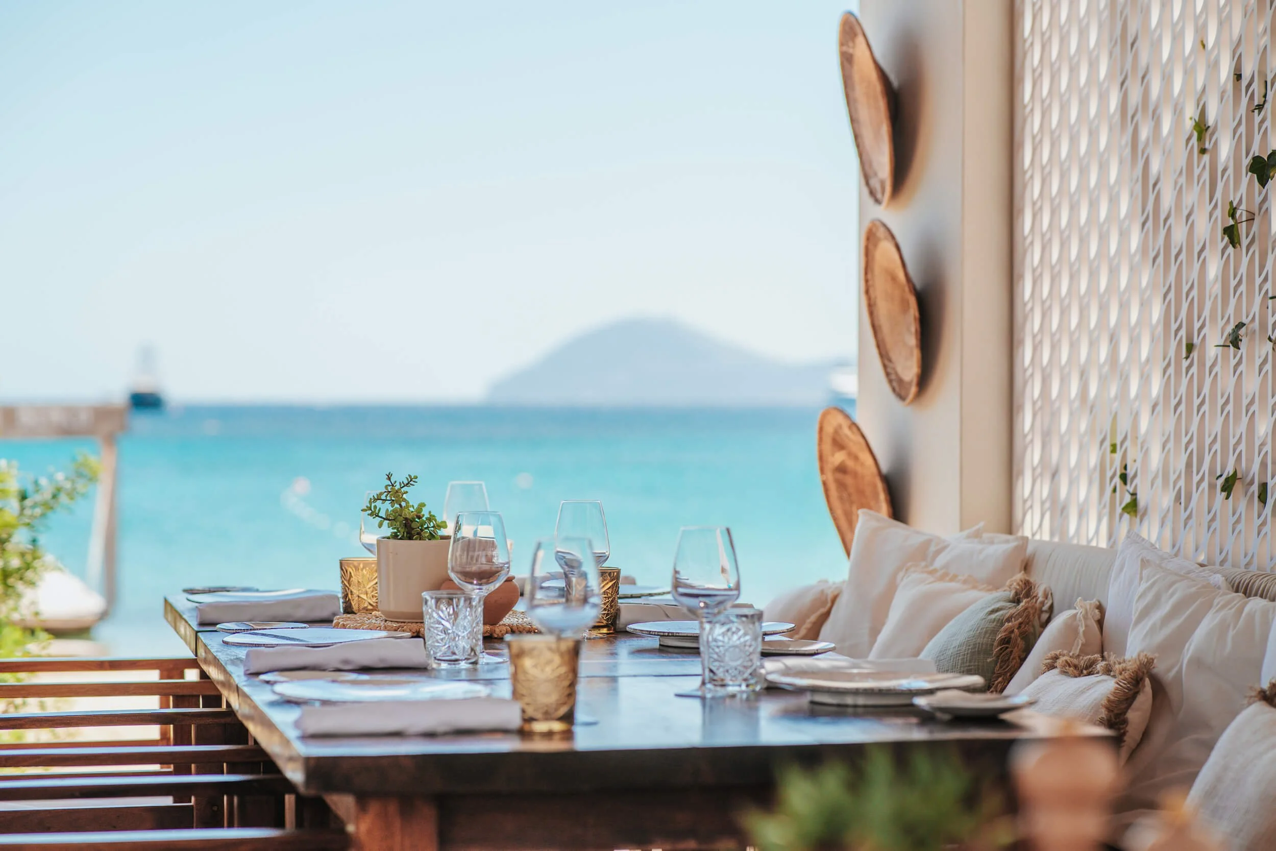 Elegant outdoor wooden table setting with wine glasses at a luxury Sardinian beach club, overlooking bright turquoise Mediterranean waters and distant coastal hills.