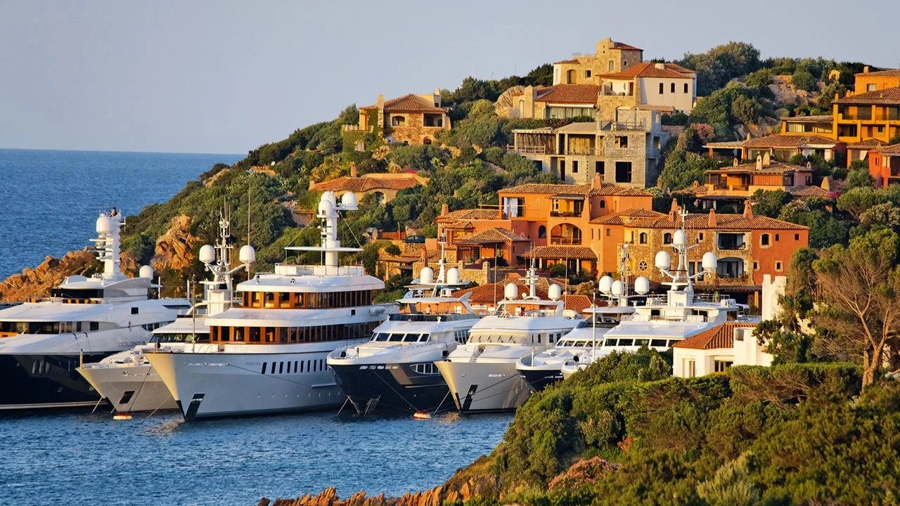 Luxury superyachts moored at Porto Cervo marina surrounded by terraced Mediterranean villas and hillsides in Costa Smeralda, Sardinia.