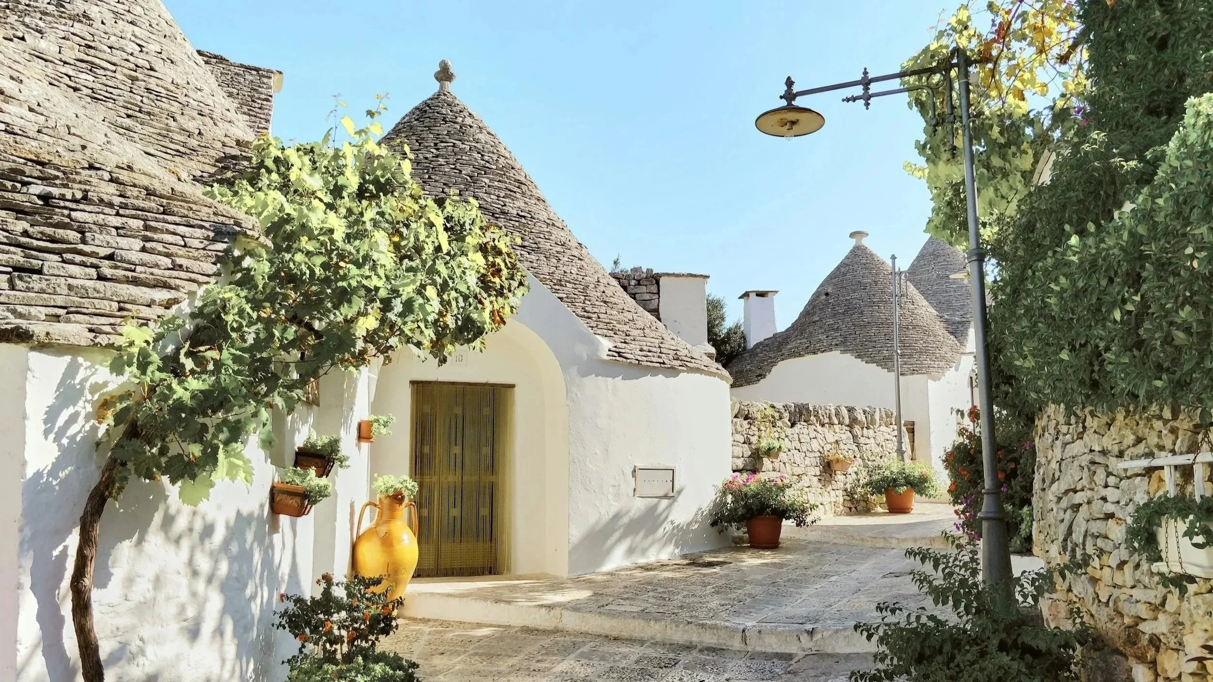 Whitewashed trulli houses with conical stone roofs lining a quiet cobblestone alley with potted plants in Alberobello, Puglia.