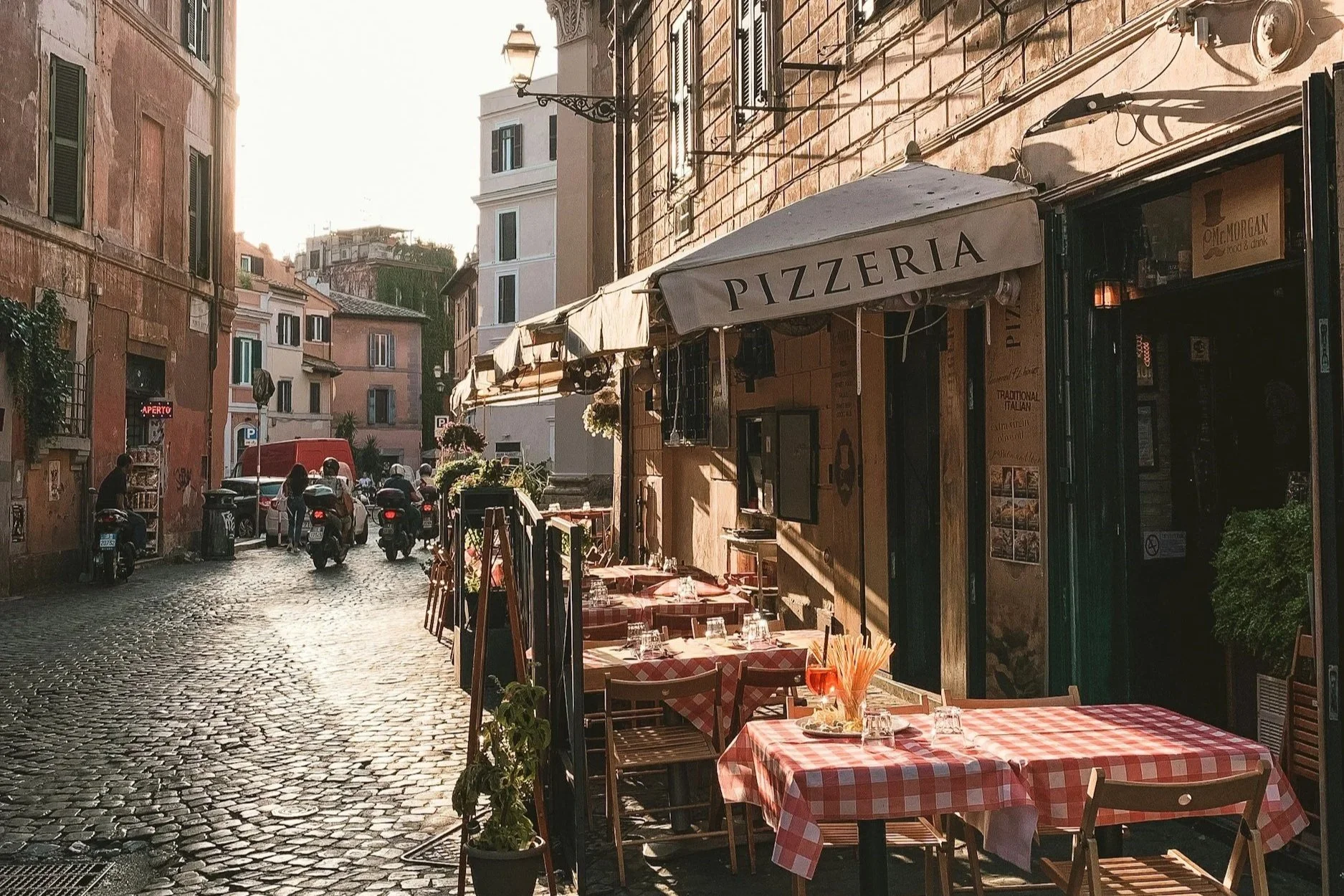 Charming cobblestone street in Rome with an outdoor pizzeria featuring red-and-white checkered tablecloths, aperitivo drinks, and warm afternoon sunlight.