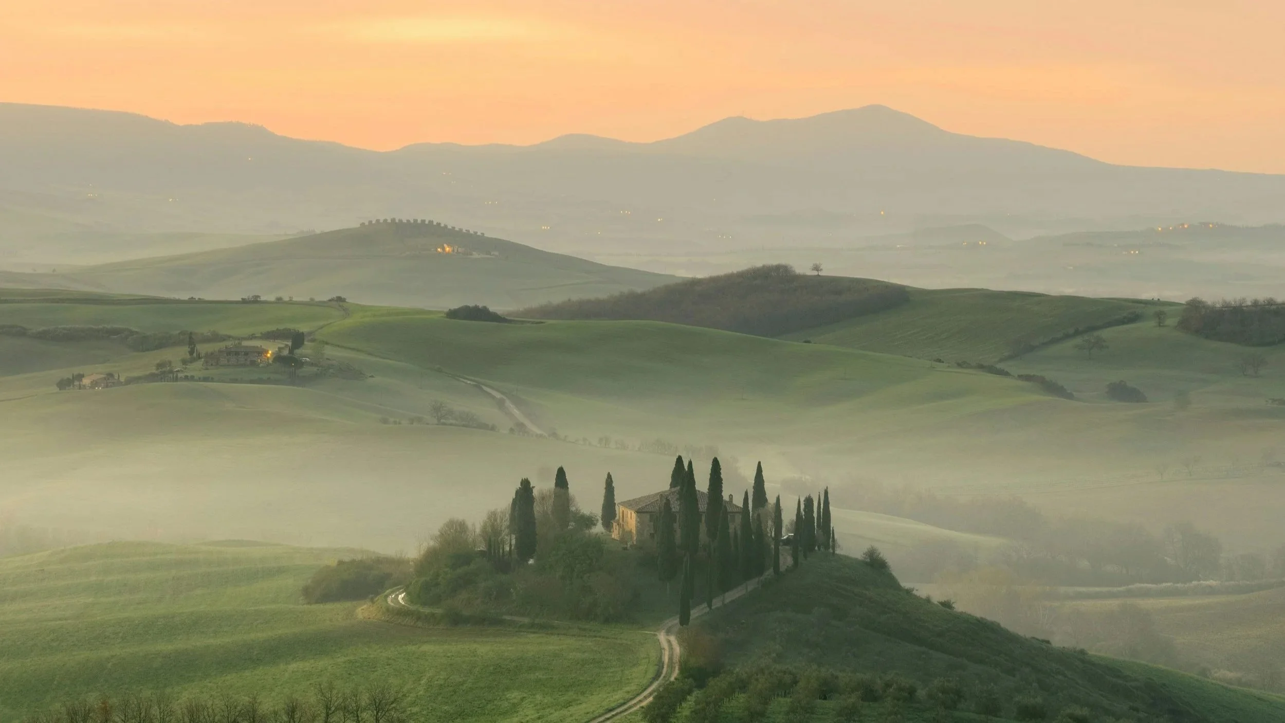 Sunrise mist rolling over green hills surrounding a traditional farmhouse framed by cypress trees in Val d'Orcia, Tuscany.