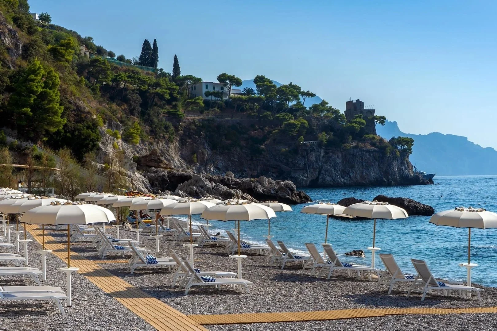 White sun umbrellas and loungers on a pebble beach along the rocky Amalfi Coast shoreline.