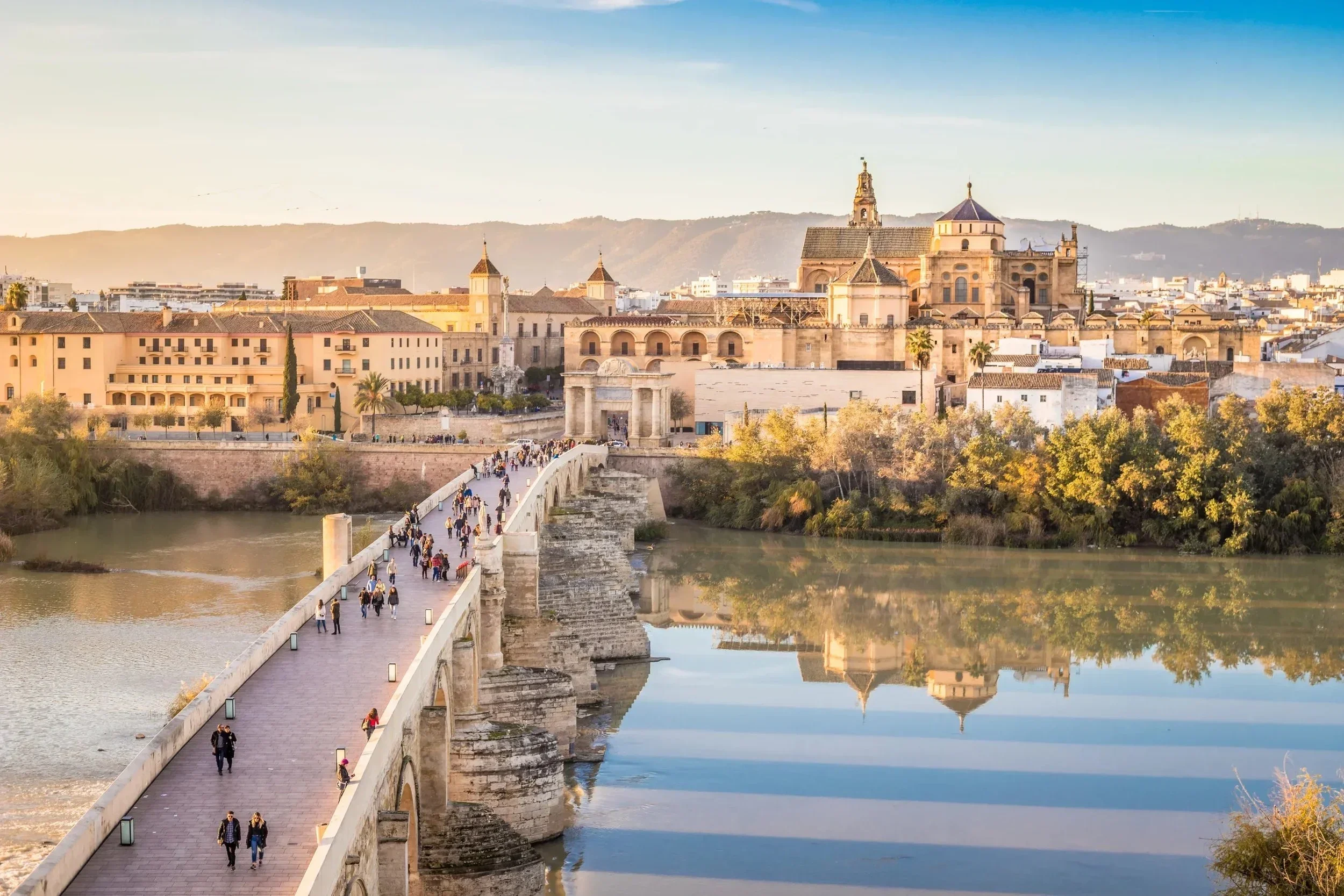 A stone bridge in Cordoba, Spain leads to a historic cityscape featuring a grand cathedral, Mosque-Cathedral Monumental Site of Córdoba. The river reflects the architecture, while people stroll under a clear blue sky.