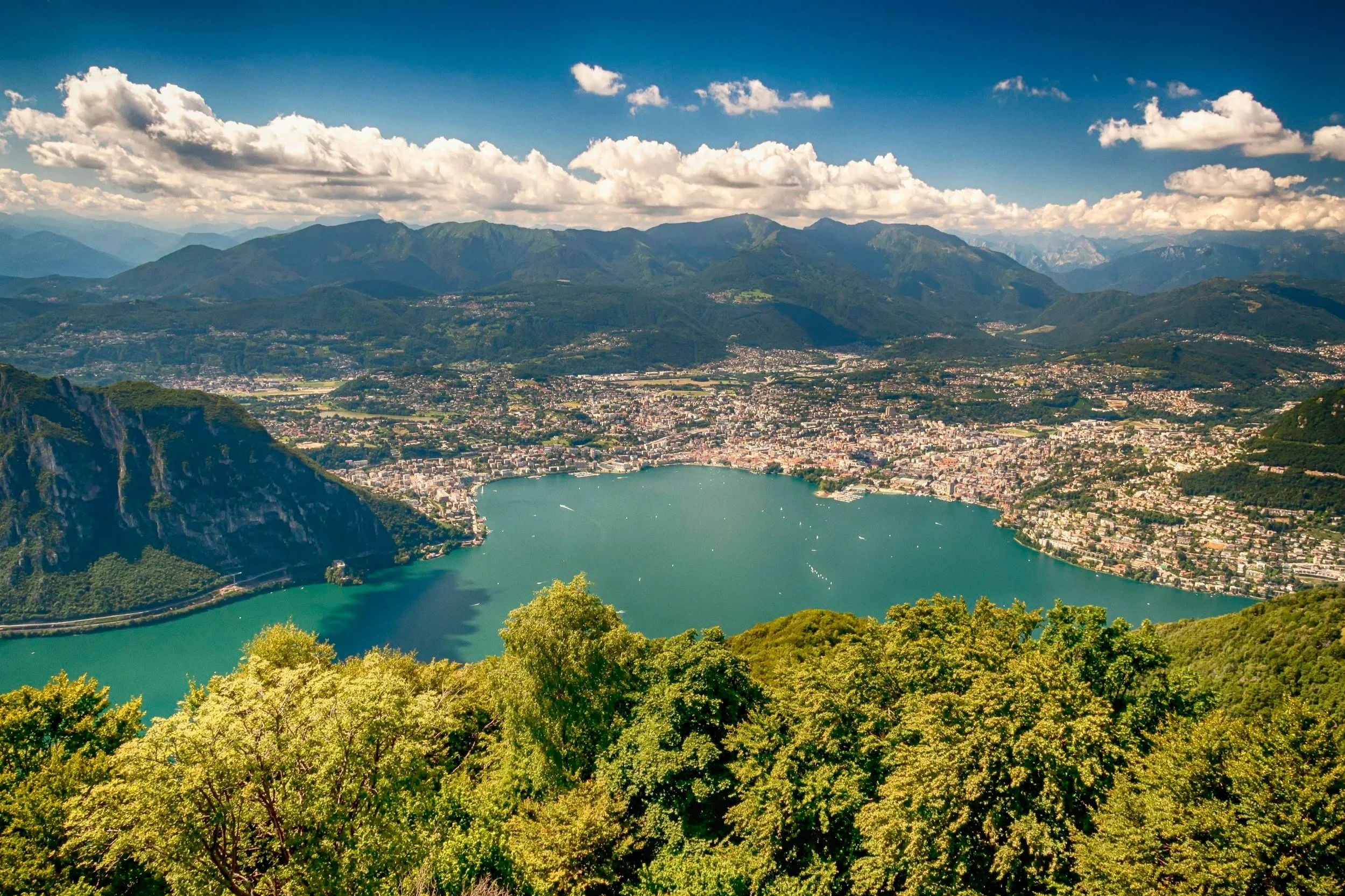 High aerial panoramic view of a deep blue alpine lake surrounded by city shorelines, green treetops, and distant mountains.