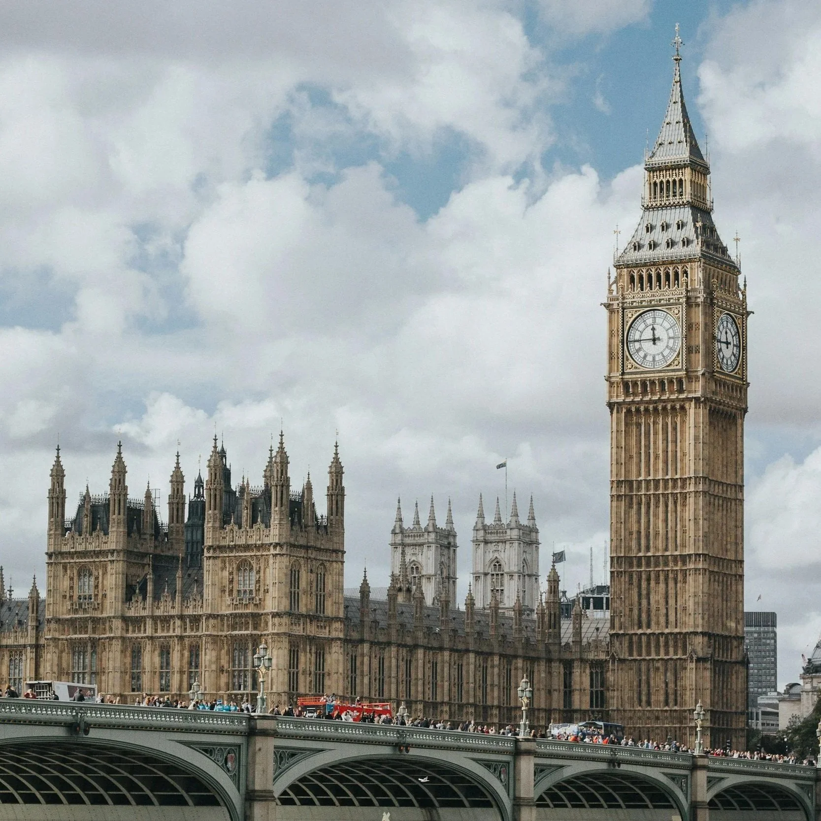 Big Ben and the Houses of Parliament in London. A cloudy sky frames the historic architecture, with red buses crossing the bridge below.