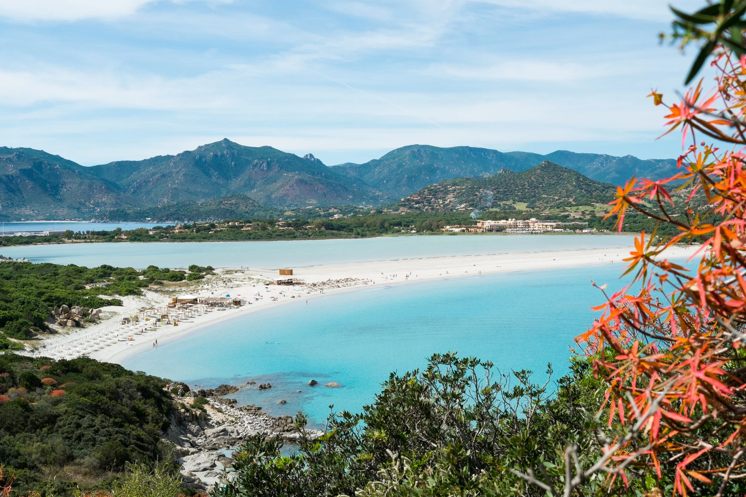 High-angle panoramic view of the crescent white sand beach at Porto Giunco separated from a calm coastal lagoon by dunes, framed by green hills in Villasimius, Sardinia.