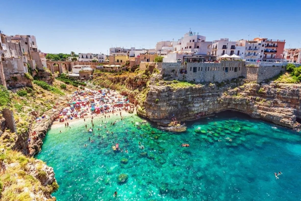Crowded pebble beach at Lama Monachile in Polignano a Mare surrounded by dramatic limestone cliffs, whitewashed buildings, and crystal-clear turquoise Adriatic waters in Puglia, Italy.