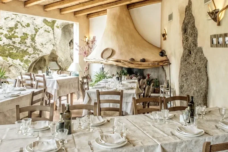 Rustic agriturismo dining room in Sardinia featuring wood-beamed ceilings, white-clothed tables, stone accents, and a large sculptural fireplace.