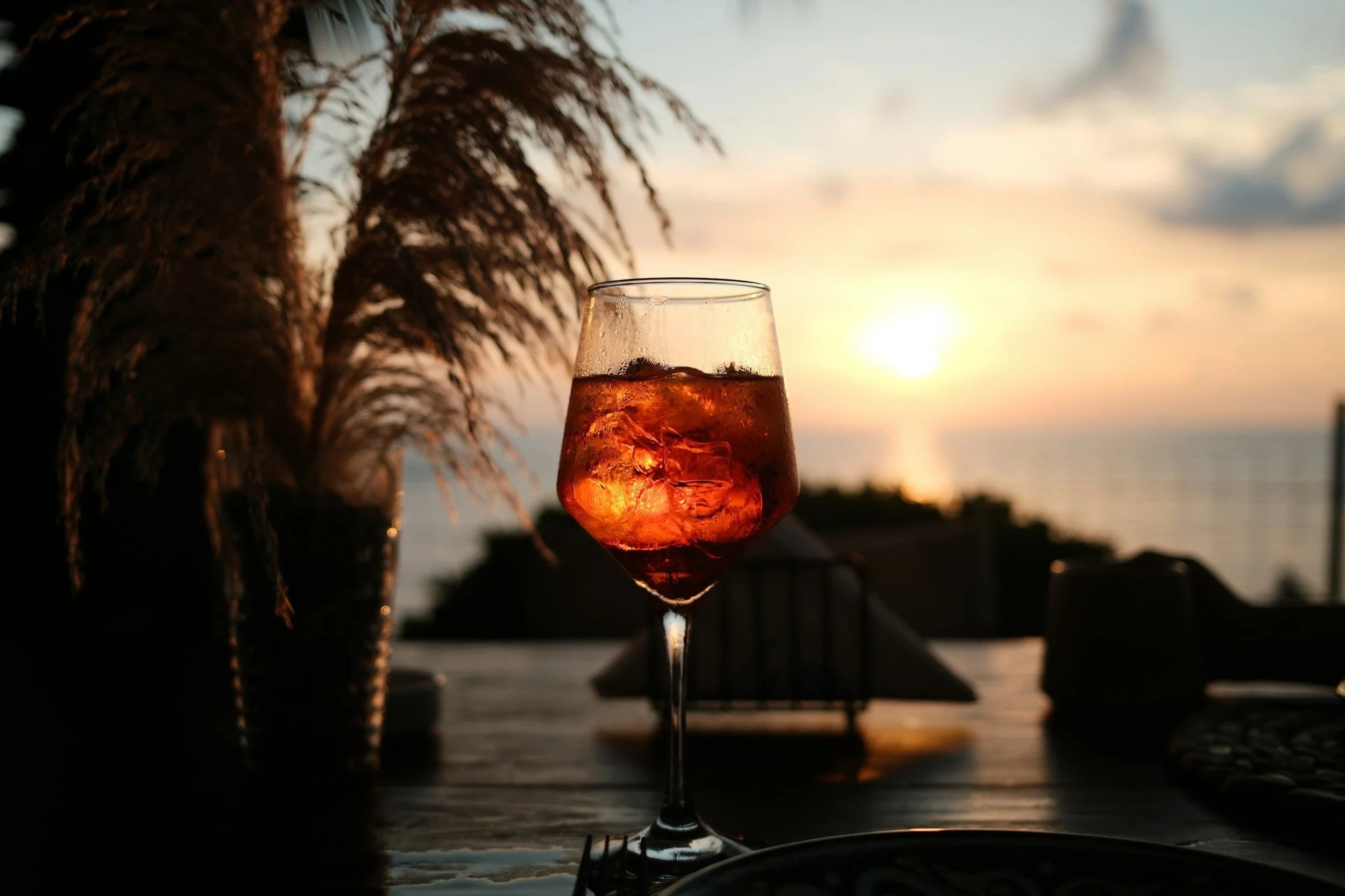 Glass of ice-cold orange Aperol Spritz cocktail on a wooden outdoor table during golden hour sunset overlooking the sea in Puglia, Italy.