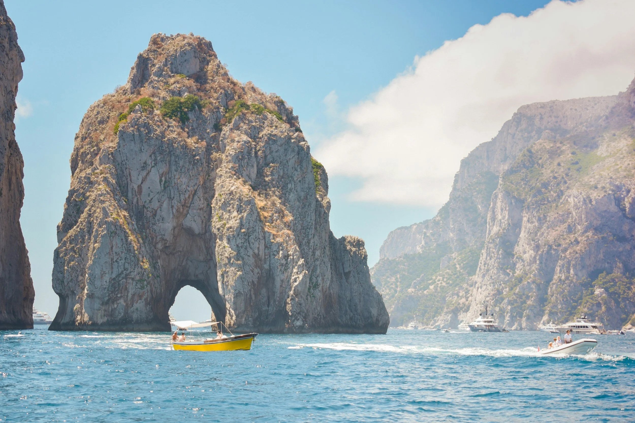 Small yellow boat passing near the Faraglioni rock formations and natural arch in Capri, Italy.