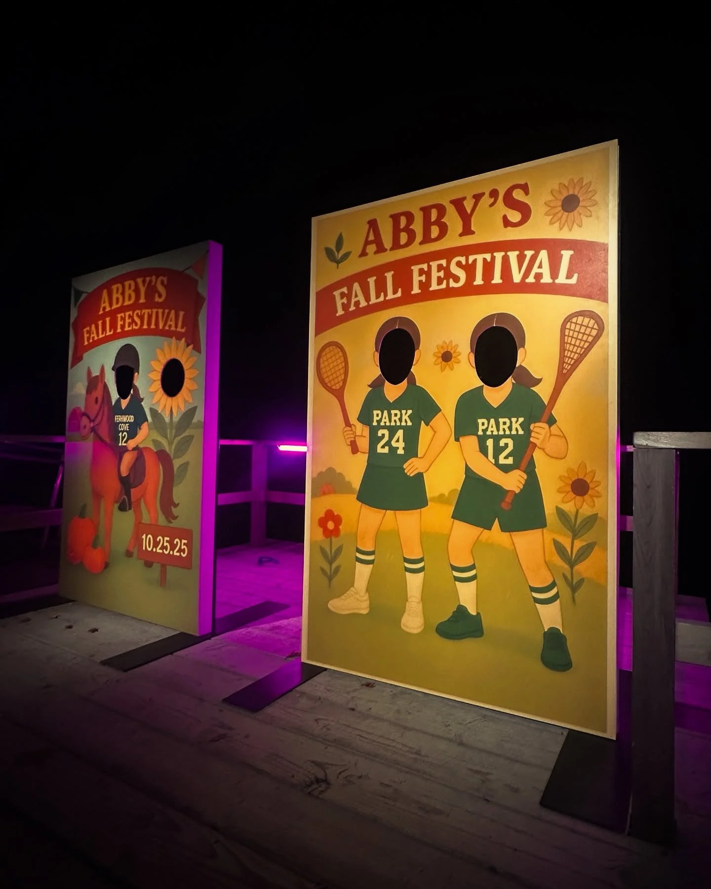 Night view of colorful outdoor festival signs for Abby's Fall Festival with cartoon illustrations of two female tennis players in green uniforms holding rackets, flowers, and pumpkins.
